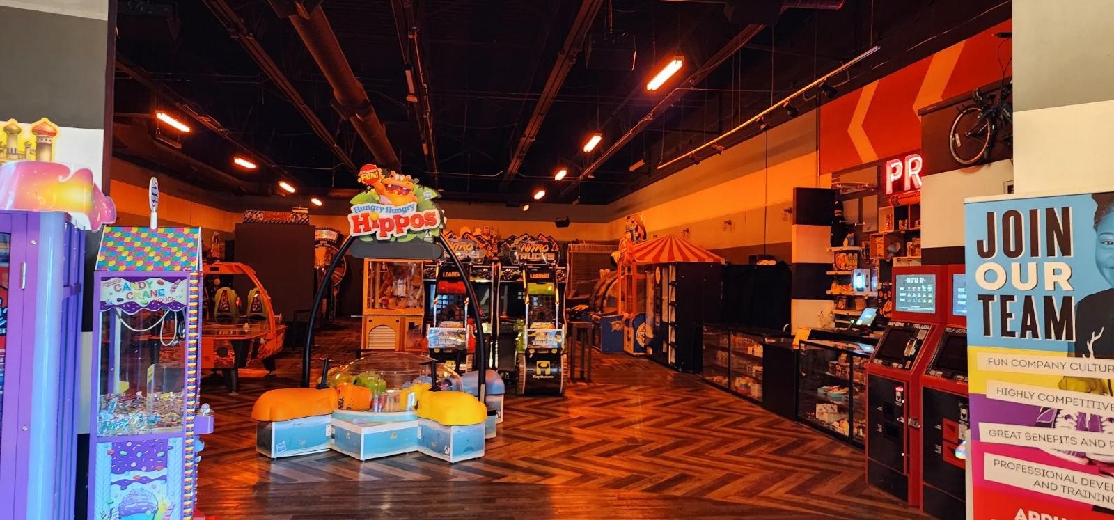 An indoor arcade and amusement center with various games and attractions, including a zone labeled 'Hungry Hungry Hippos,' and a sign inviting people to join the team.