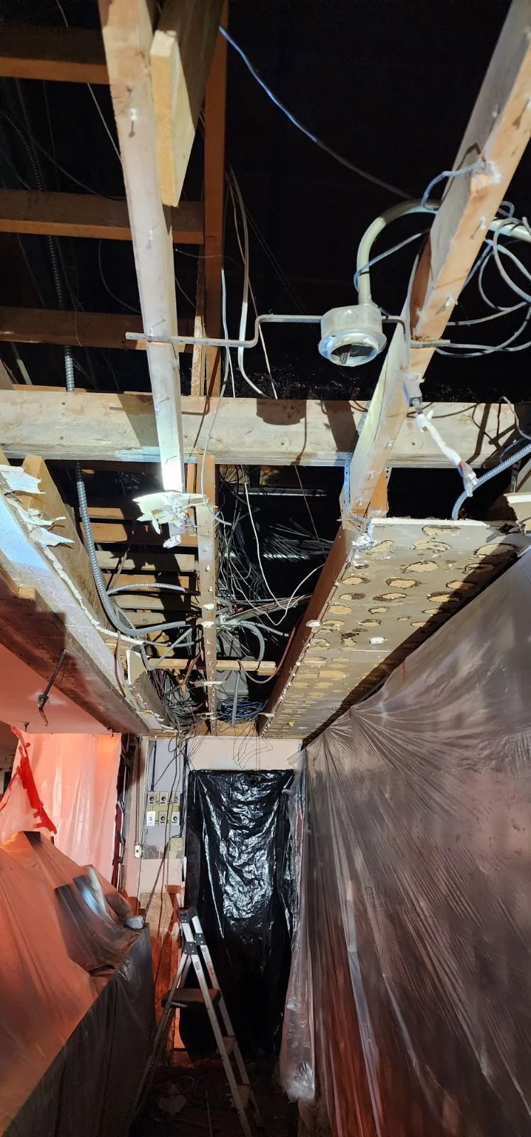View of exposed ceiling with electrical wiring and studs, plastic sheeting covering walls, and a stepladder in a construction or renovation site.