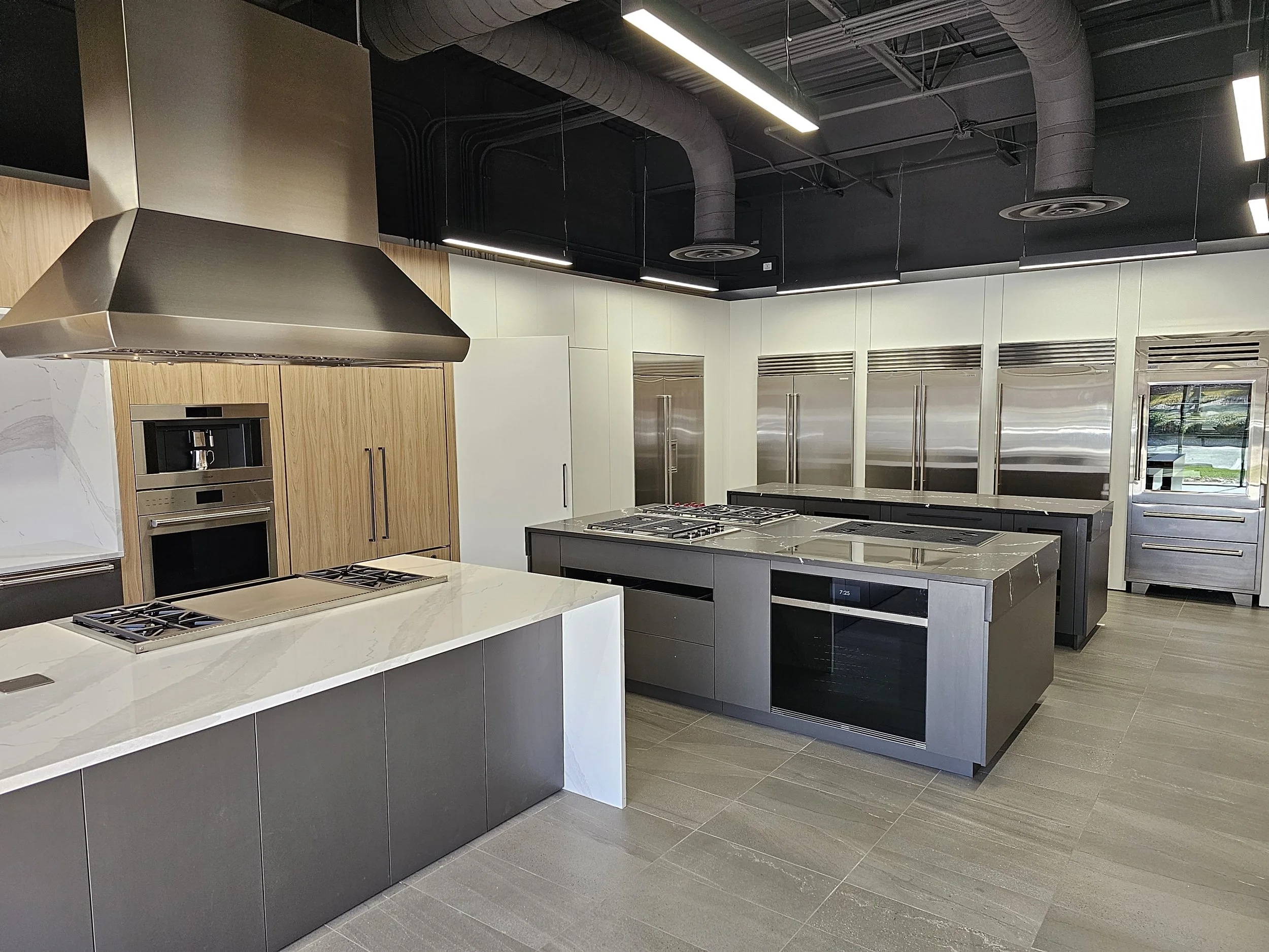 Modern kitchen with stainless steel appliances, black and grey cabinets, marble countertops, and exposed industrial ceiling pipes.