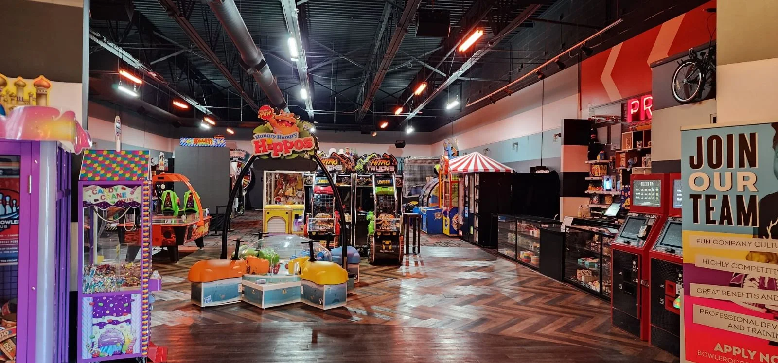Interior of an arcade filled with various game machines and amusement rides, including a large funhouse sign, a bubble gum machine, and arcade cabinets, with a wooden floor and colorful decor.