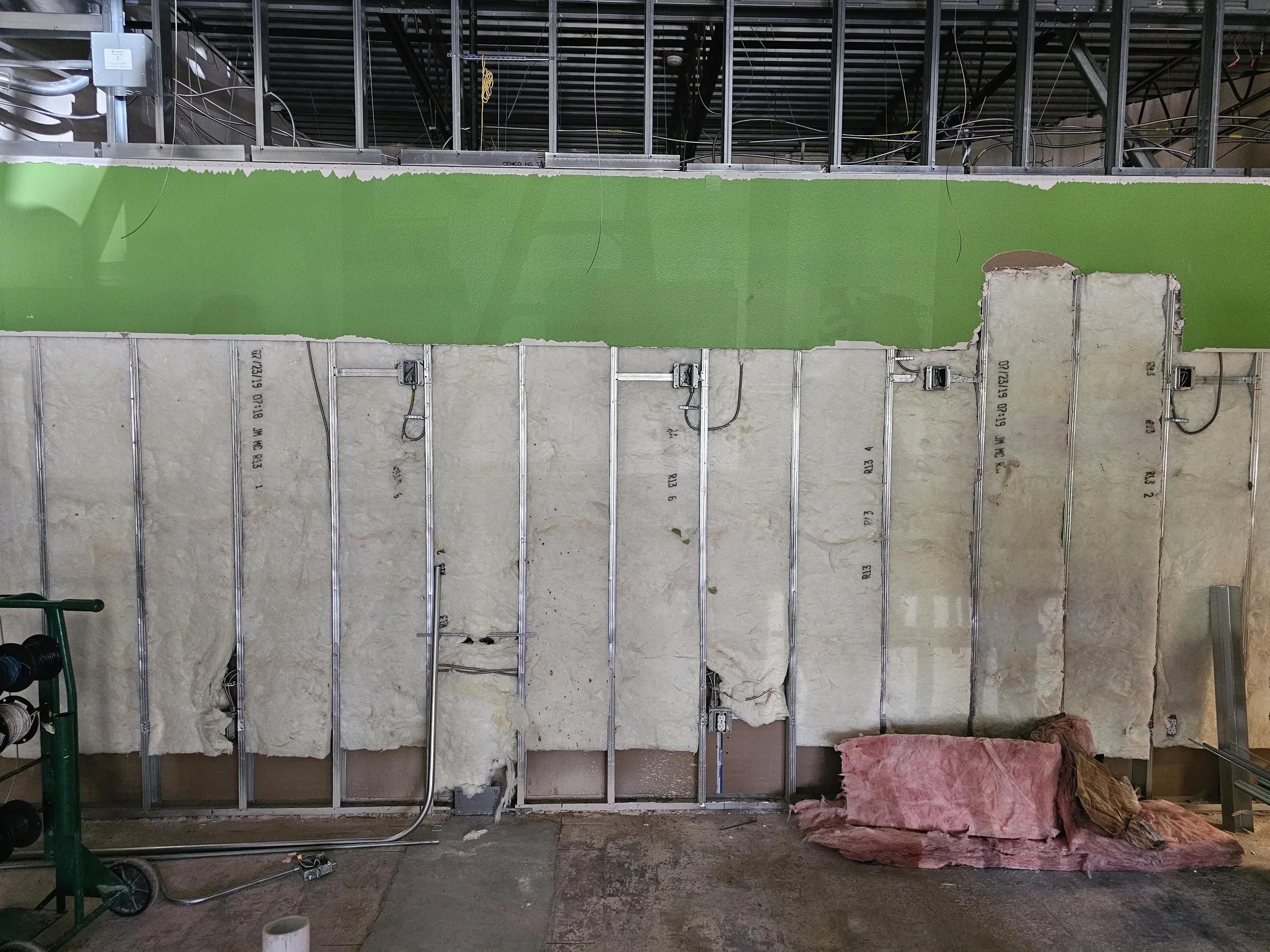Interior view of a building under construction, showing metal framing for walls, insulation on the lower walls, and a partially finished green painted section of the wall. Construction tools and materials are visible on the floor.