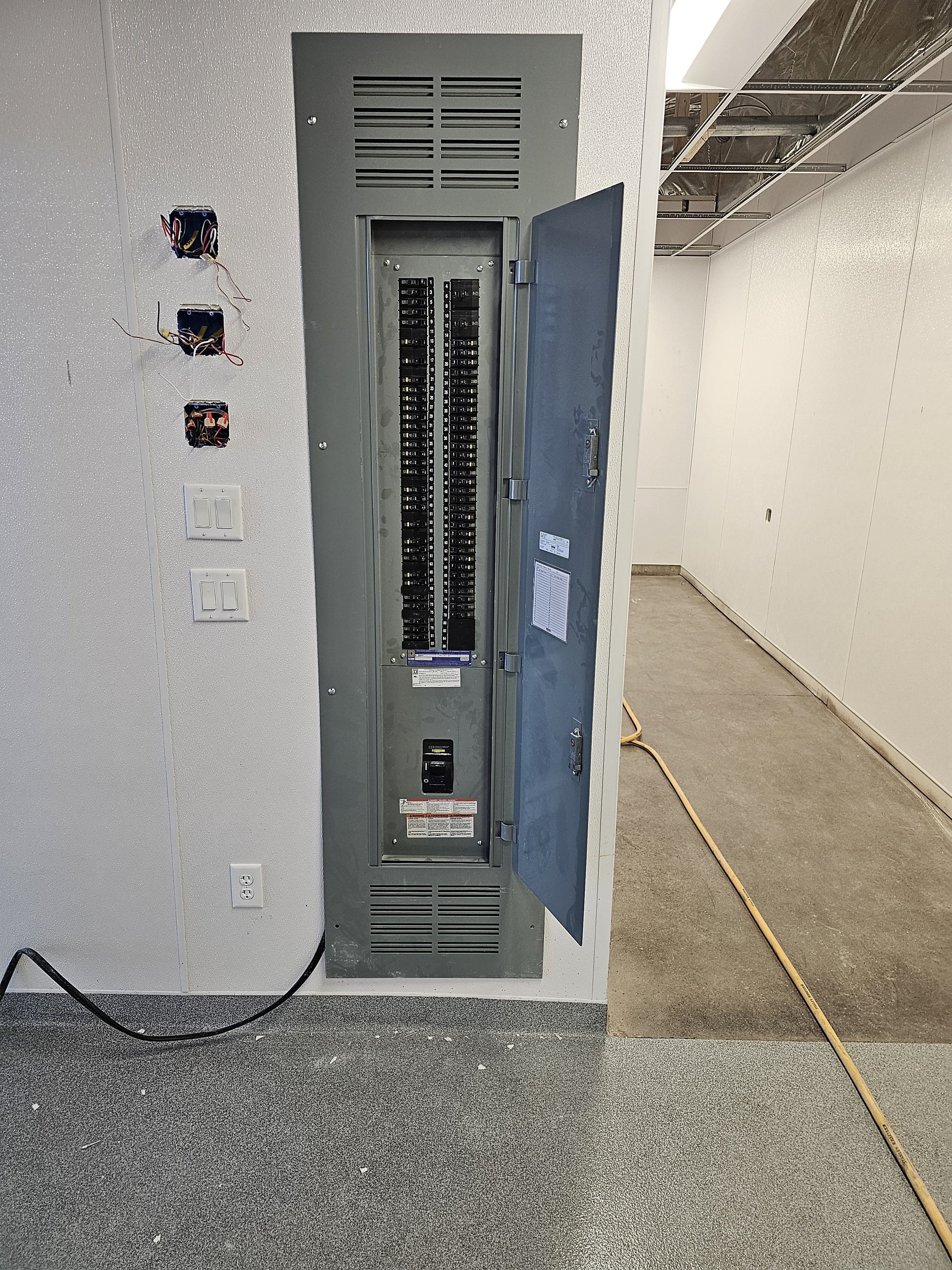 Interior of an electrical or telecommunications panel box with an open door, showing various circuit breakers inside. Three electrical wire boxes are mounted on the wall next to the panel, with wires protruding from them. There are also electrical ou