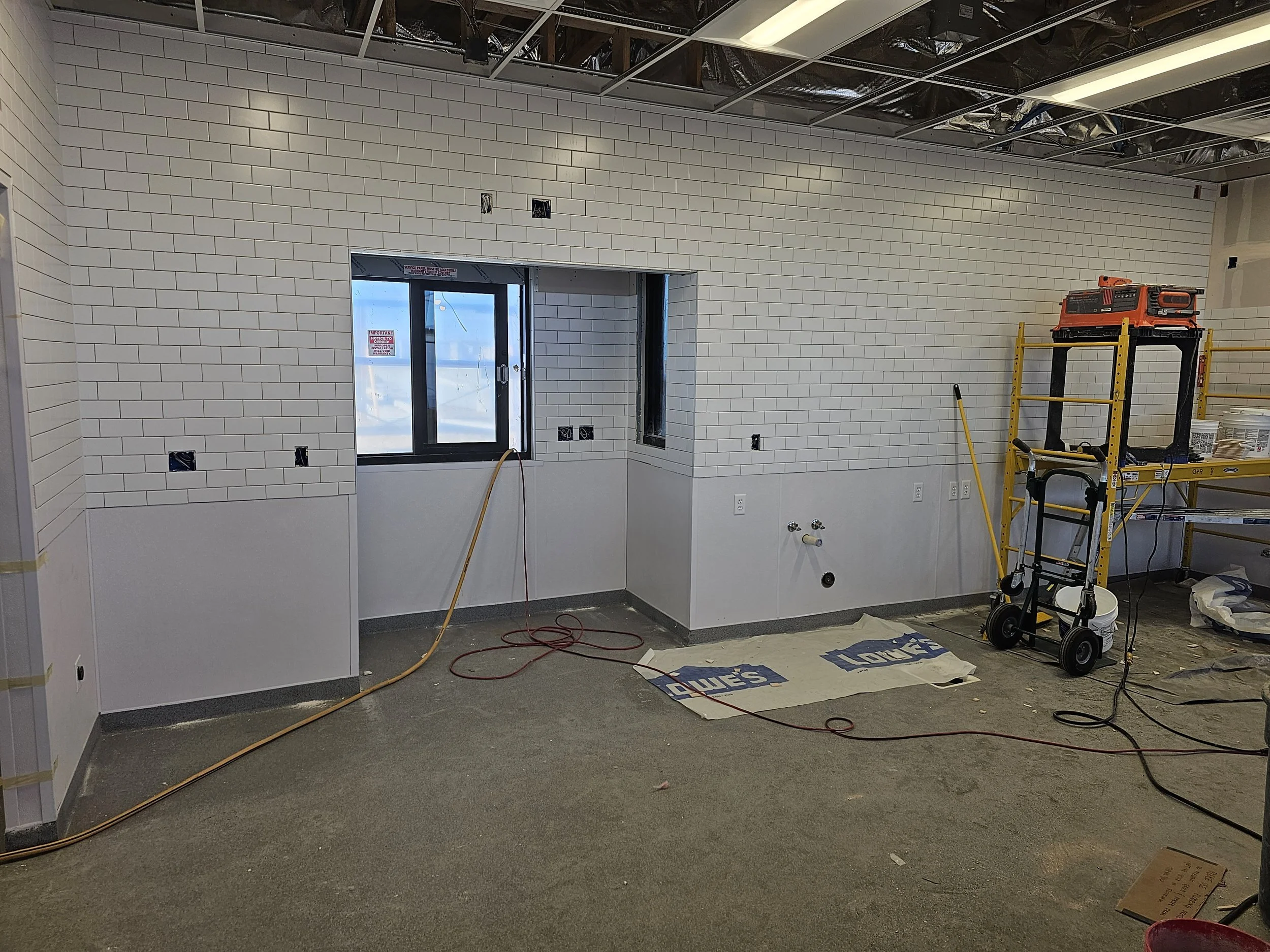 Interior of a room under construction with tiled walls, open ceiling exposing insulation and metal framing, a window, a yellow rolling scaffold, some tools, and construction materials on the floor.