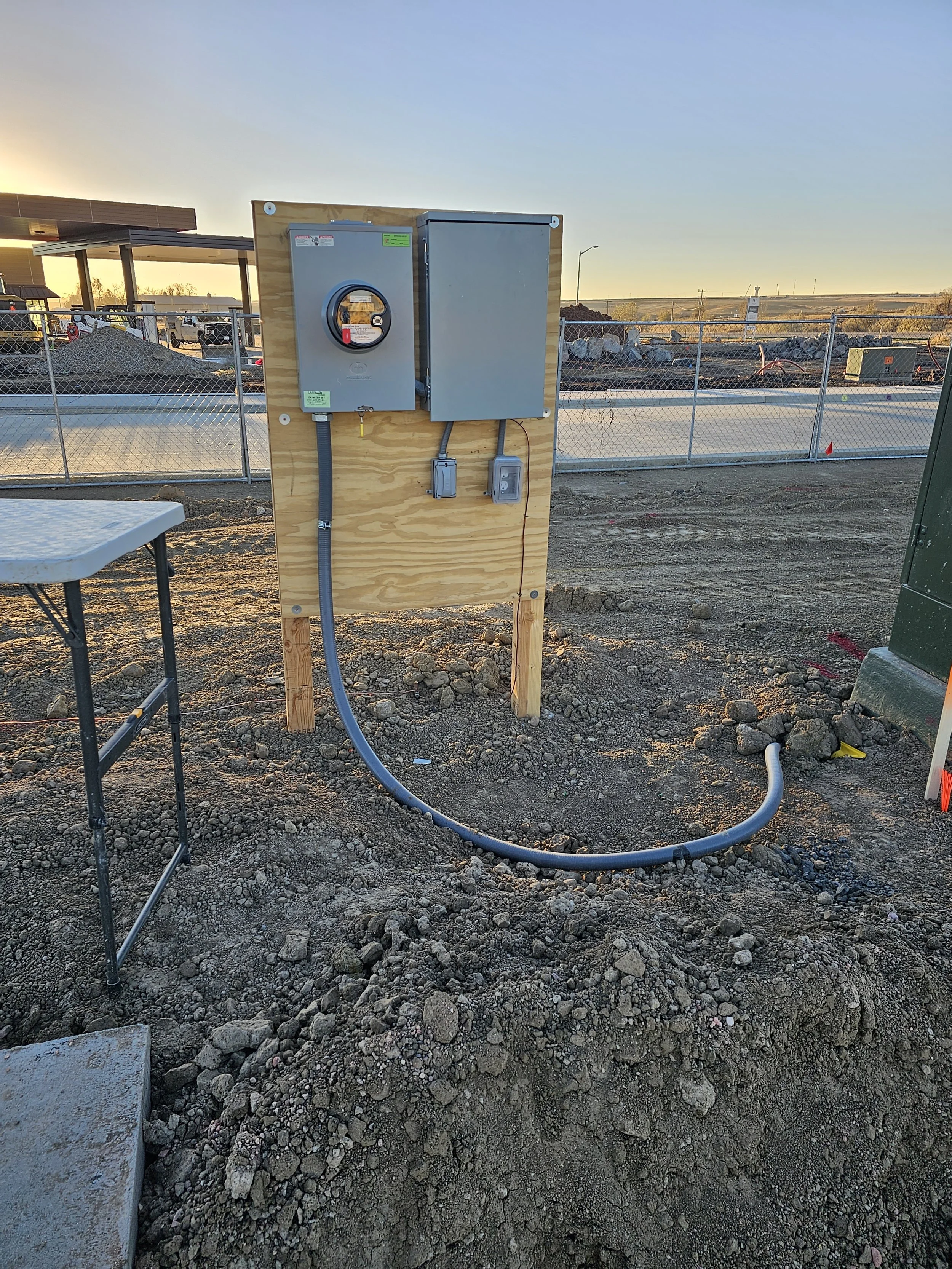 Outdoor construction site with electrical panels mounted on a wooden post, surrounded by dirt and gravel, with a chain-link fence and open landscape in the background during sunset.