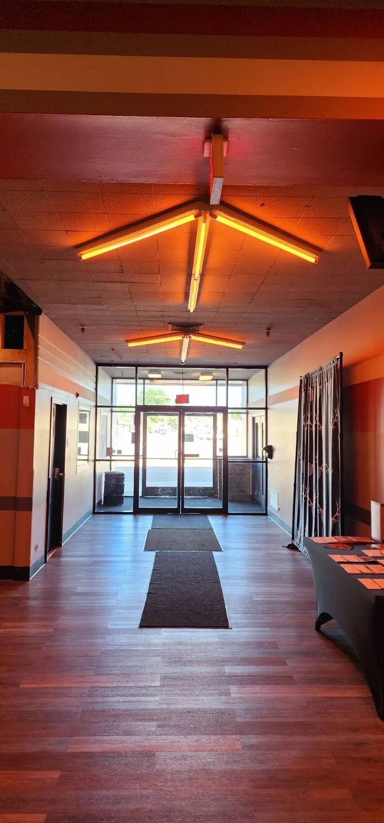 Entrance hallway with wood flooring, illuminated by modern orange lighting fixtures on the ceiling, leading to glass doors and windows with an outside view.