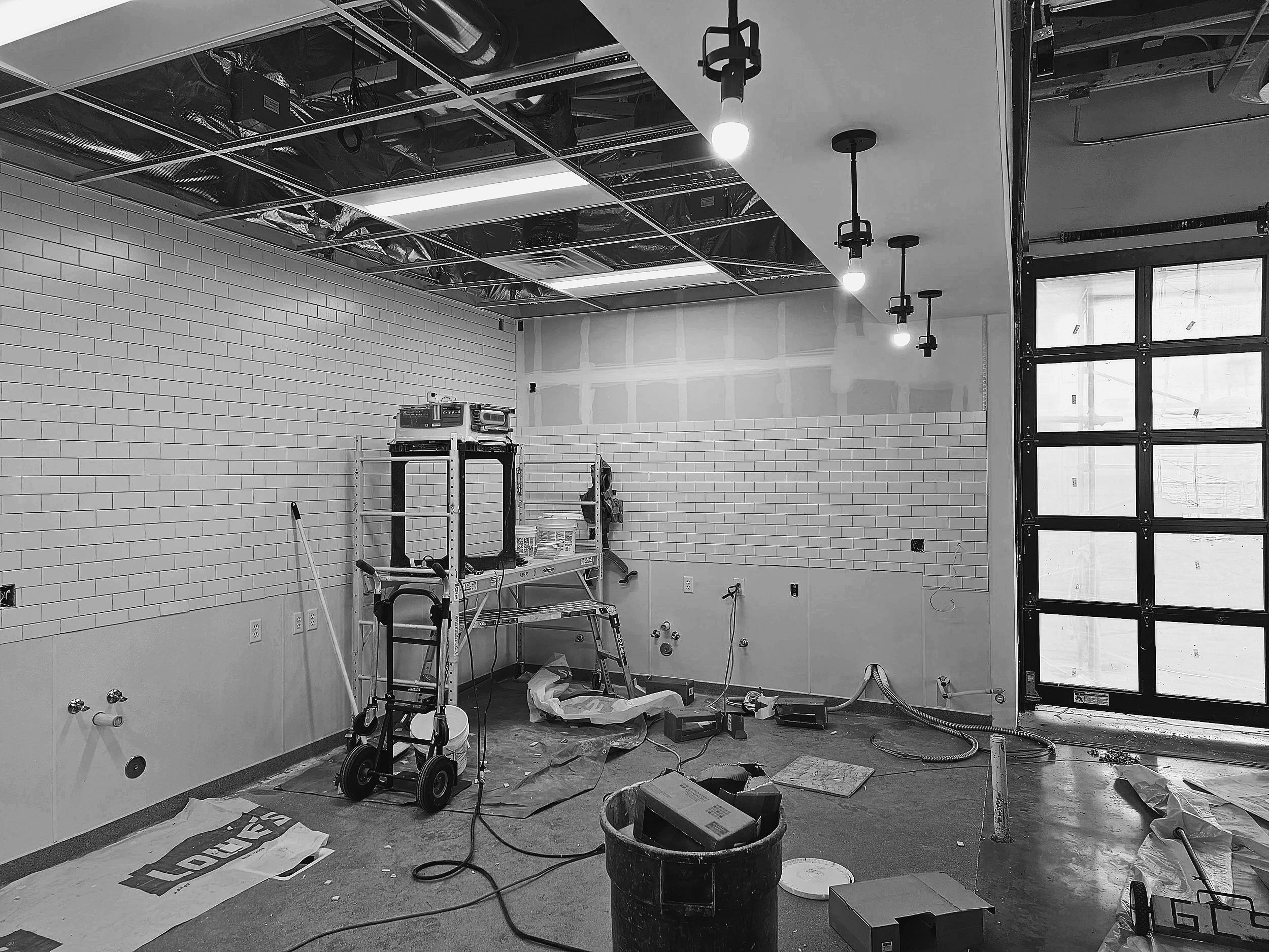 Under construction room with exposed ceiling and light fixtures, brick walls, construction tools, and materials scattered on the floor.