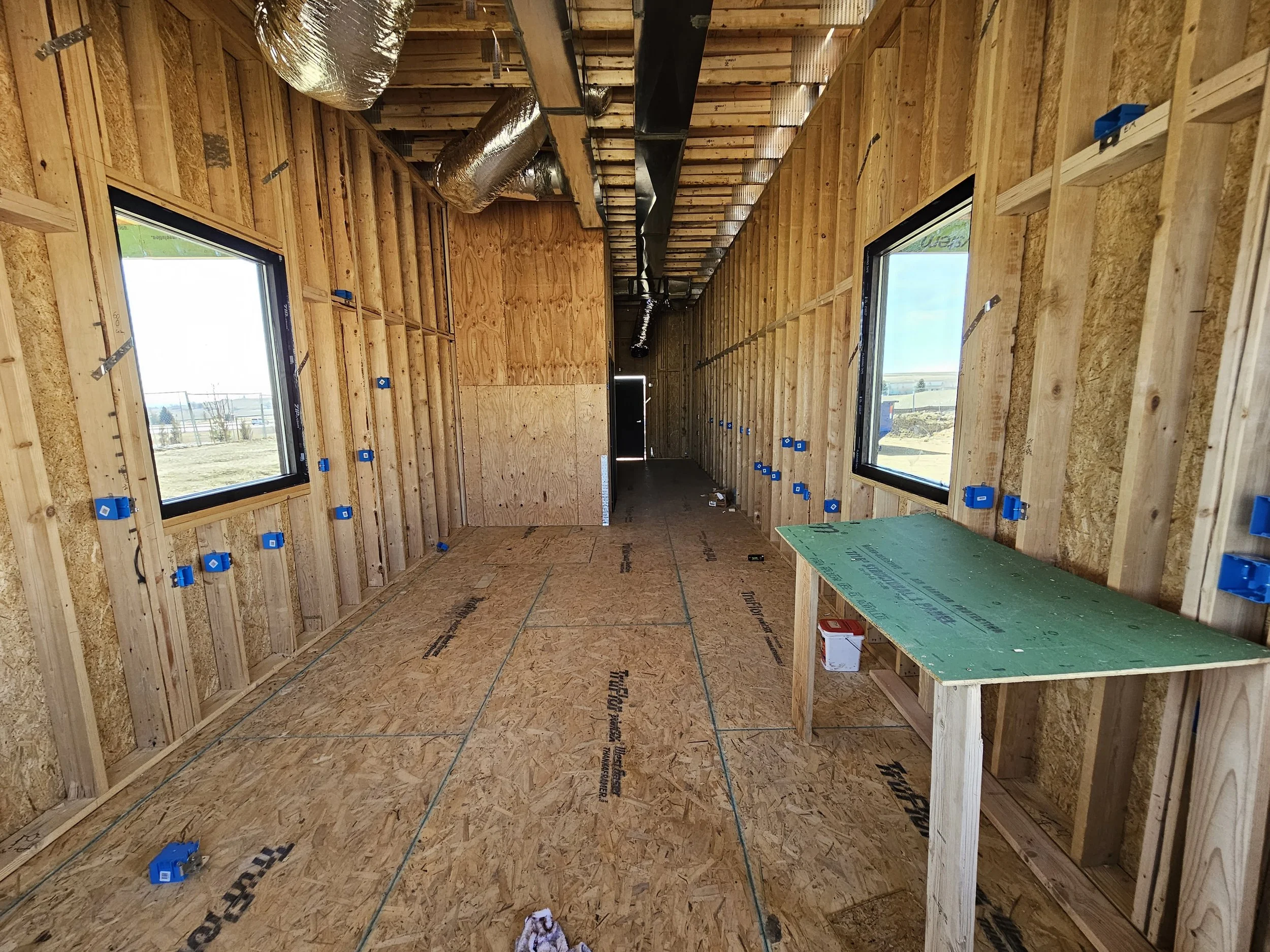 Interior view of a building under construction with exposed wooden framing, insulation ductwork hanging from the ceiling, and blue electrical box mounts on the walls. Two large windows show a sunny outdoor scene.