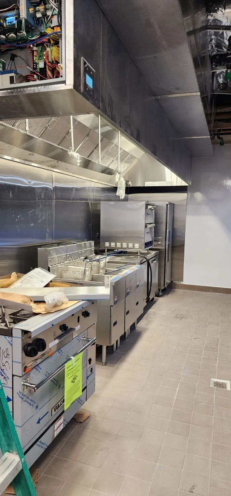 Commercial kitchen with stainless steel appliances, including a deep fryer, refrigerator, and ventilation hood, with some utensils and equipment on the countertops.