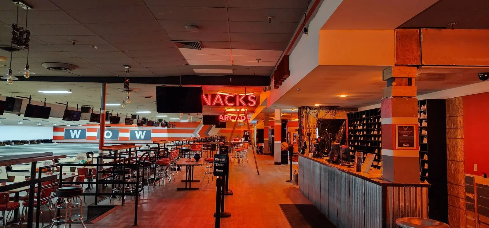 Interior of a bowling alley with seating area, multiple screens, and neon signs reading 'SNACKS' and 'ARCADE'. The lighting is dim with a warm, orange glow.
