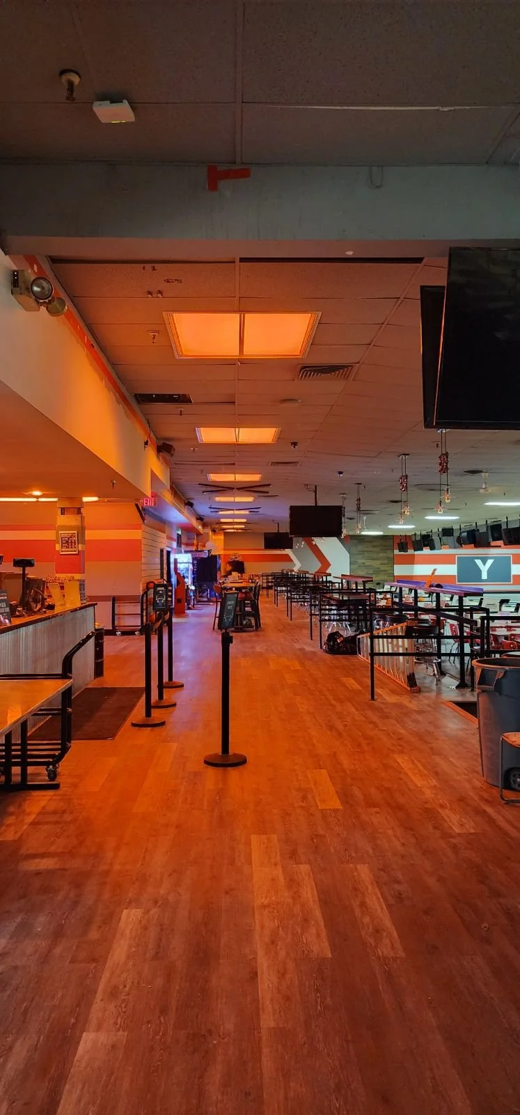 Empty indoor sports or entertainment venue with wooden flooring, display screens, and bar-height tables. Roped-off areas suggesting a line or check-in queue. Dim lighting with orange accents.