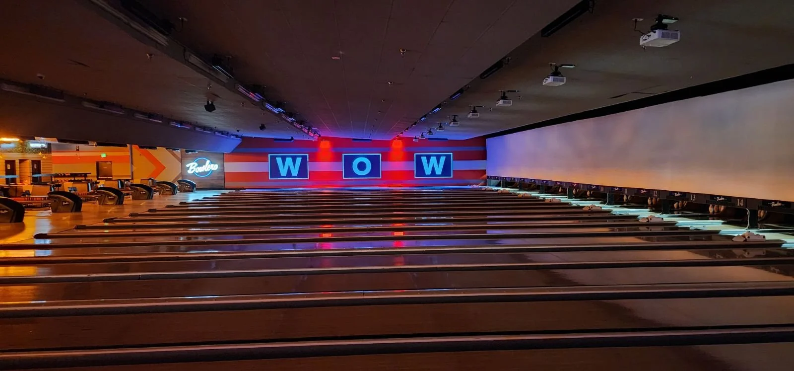 Empty bowling alley with lanes, pins, and a neon sign that says 'Bowlero' on the back wall, along with illuminated letters spelling 'WOW' on the LED screens.