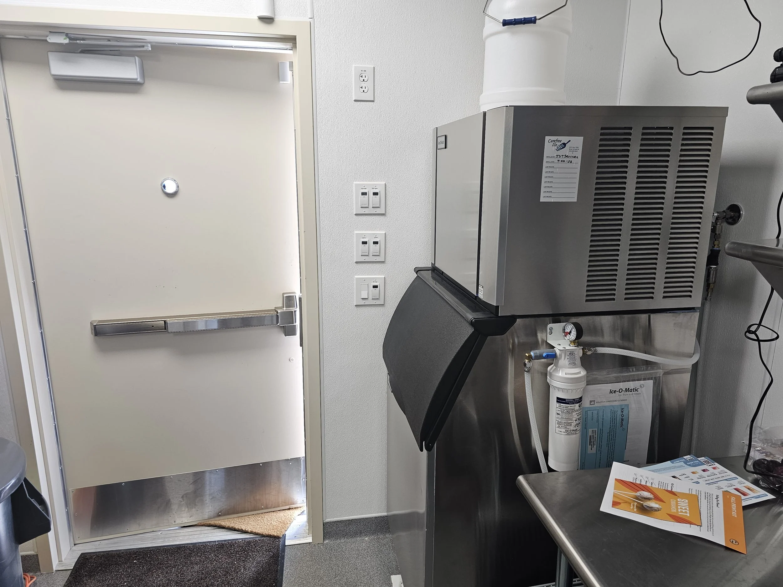 A small room with a closed white door, a wall-mounted electrical outlet, and a countertop with a countertop ice machine. The door has a metal push bar and a small round peephole. The ice machine is metallic, with a white water bottle on top and a lab