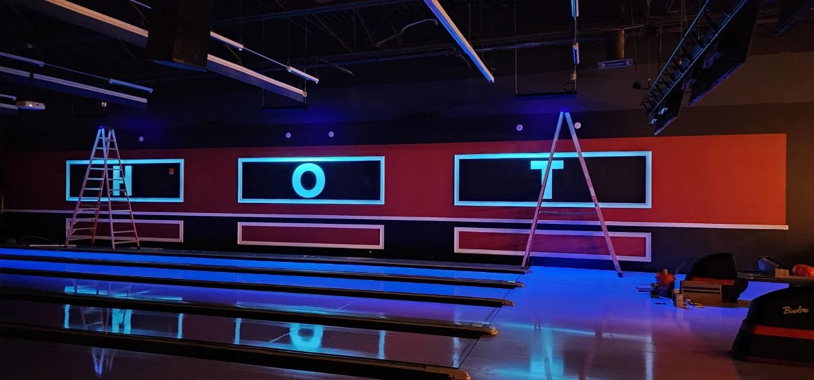 Empty bowling alley with lanes, ladder, and illuminated wall with the letters 'O', 'T', and 'O' in neon lights.