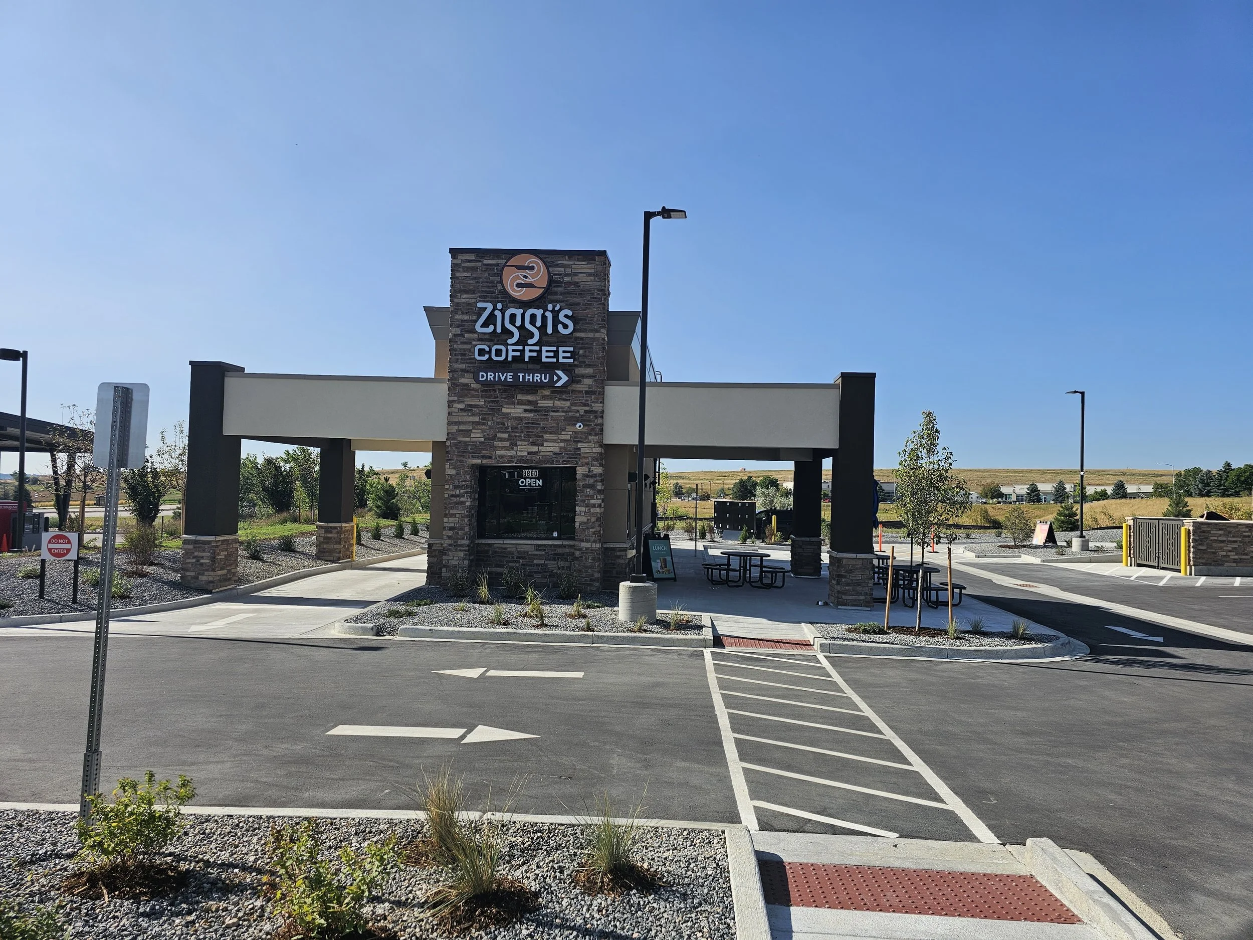 Empty parking lot outside a coffee shop named Ziggi's Coffee with drive-thru, with a blue sky and some trees and buildings in the background.