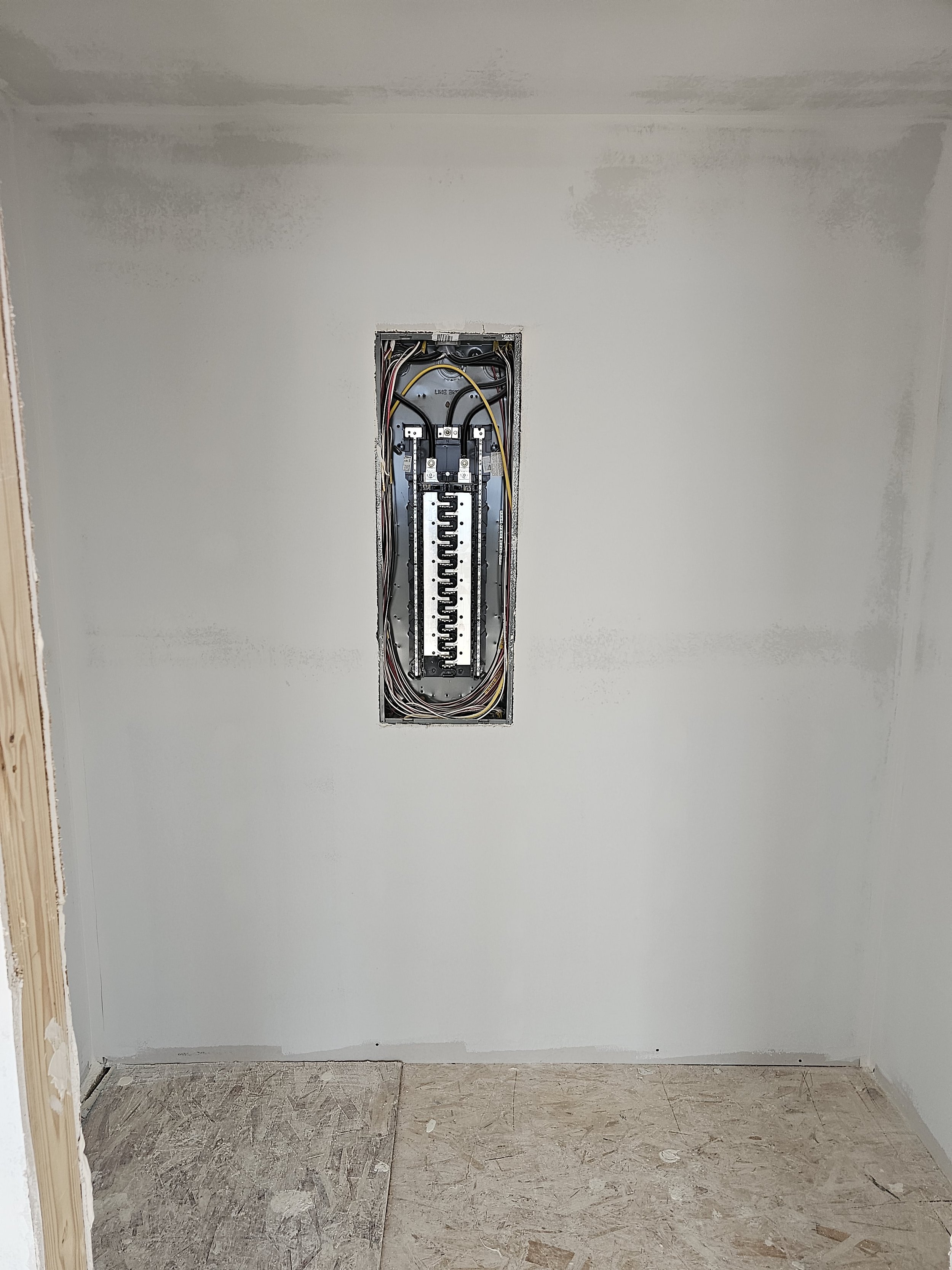 An unfinished room with drywall installed, with an electrical box in the wall containing wires, and a partially finished floor with plywood and some debris.