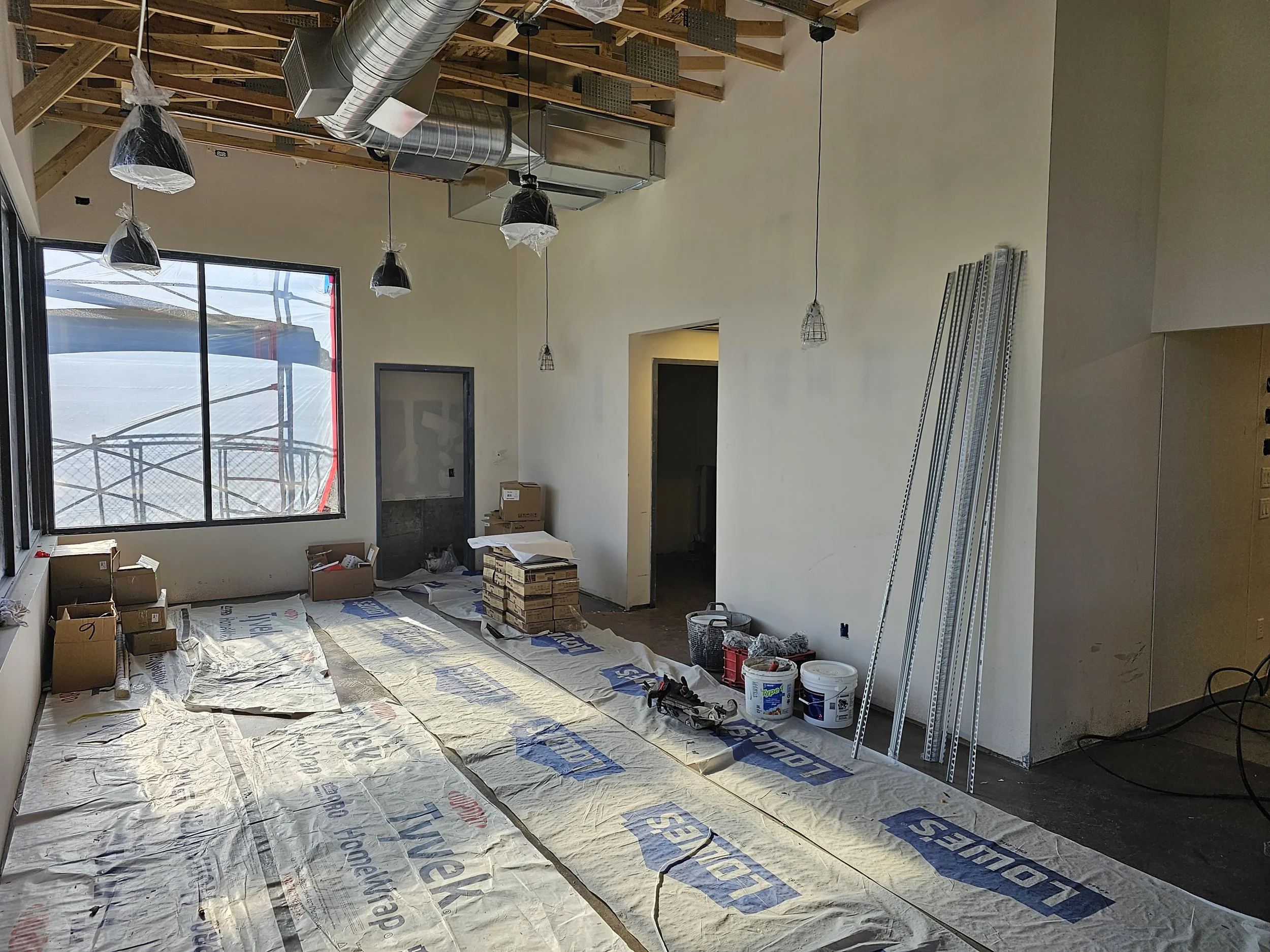 Interior of a building under construction or renovation with exposed wooden roof trusses, vents, hanging light fixtures, large window, boxes, construction materials, and protective covering on the floor.