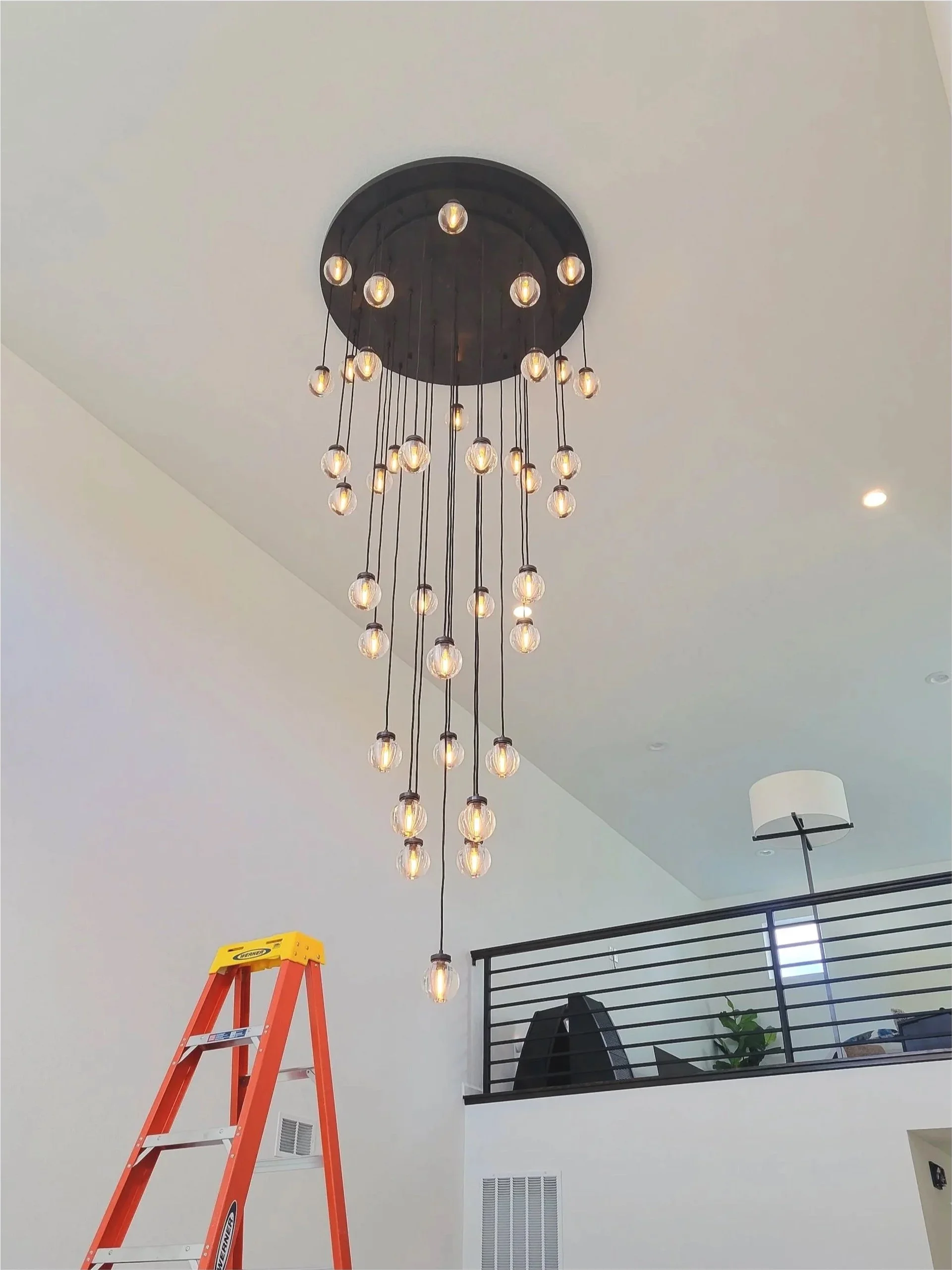 Modern chandelier with multiple hanging bulbs in a spacious room, with a black railing on upper level and a red step ladder in the foreground.