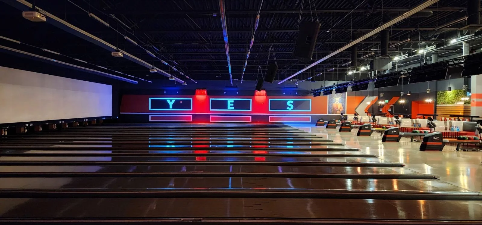 A modern indoor bowling alley with multiple lanes, neon signs spelling Y E S on the back wall, and seating areas on the right.