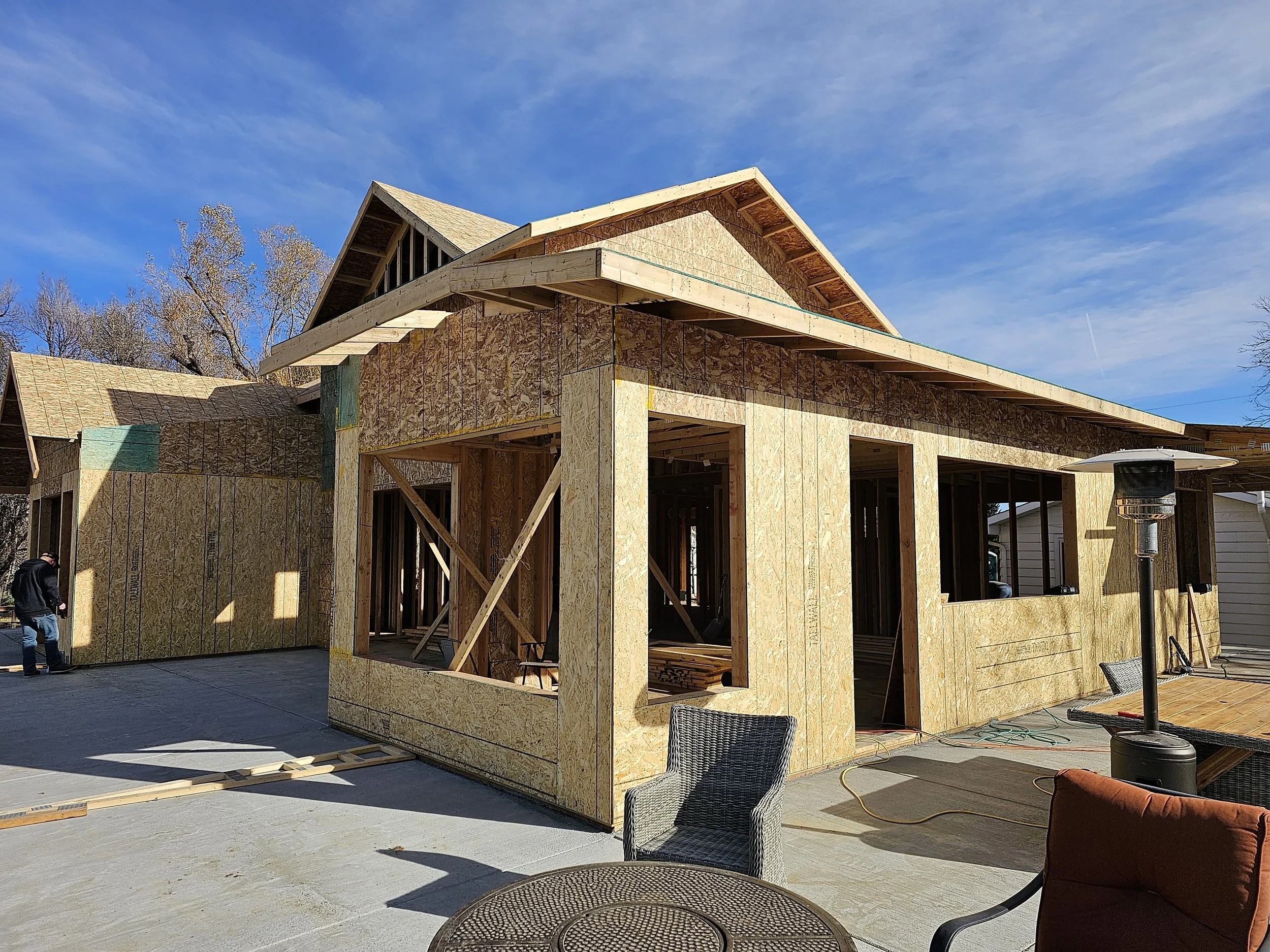 Construction site of a house with wooden framing and sheathing, under a blue sky with some clouds, and outdoor chairs in the foreground.