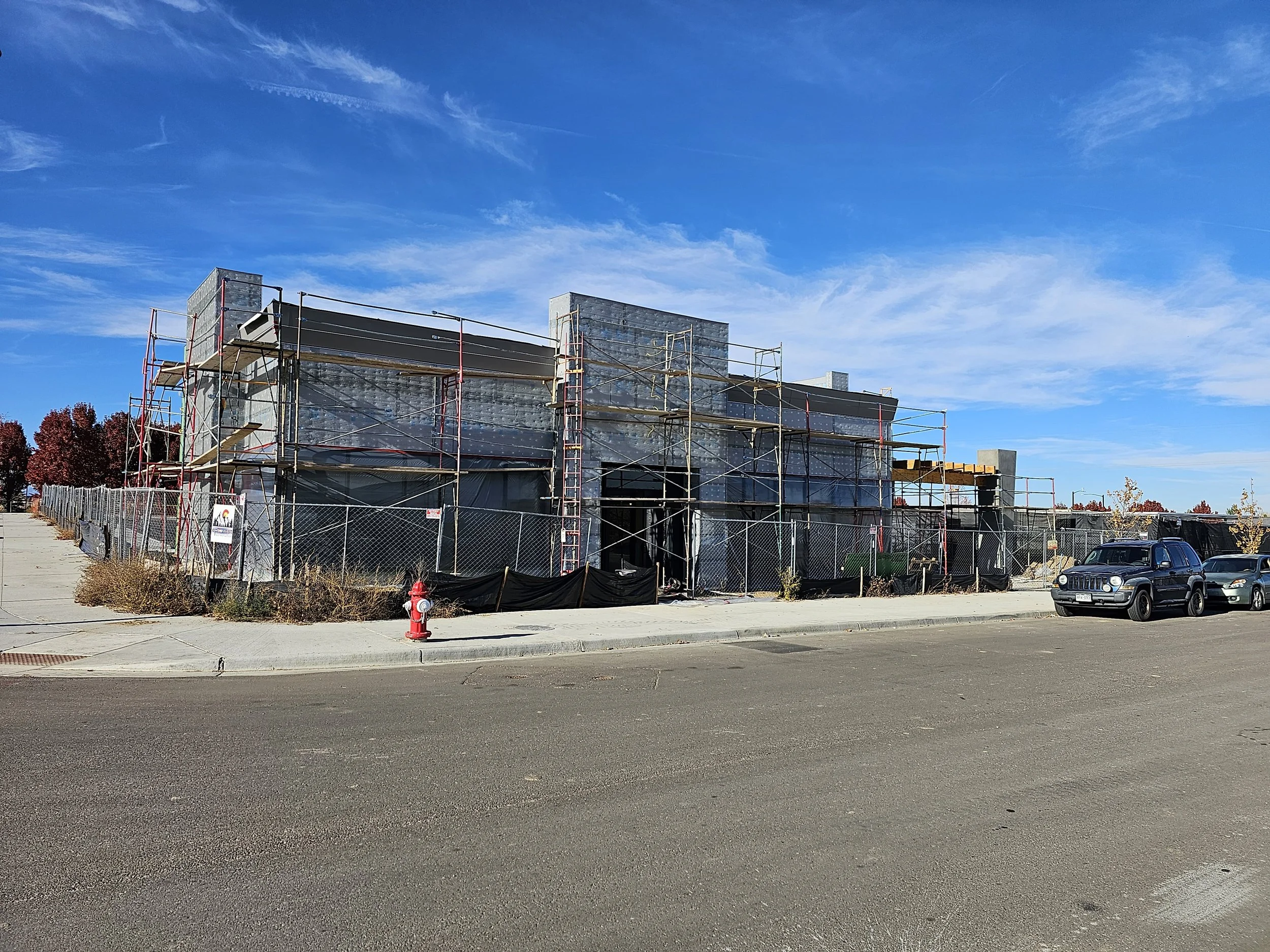 Construction site with a modern building under construction, surrounded by scaffolding, with a clear blue sky overhead and a row of trees in the background.