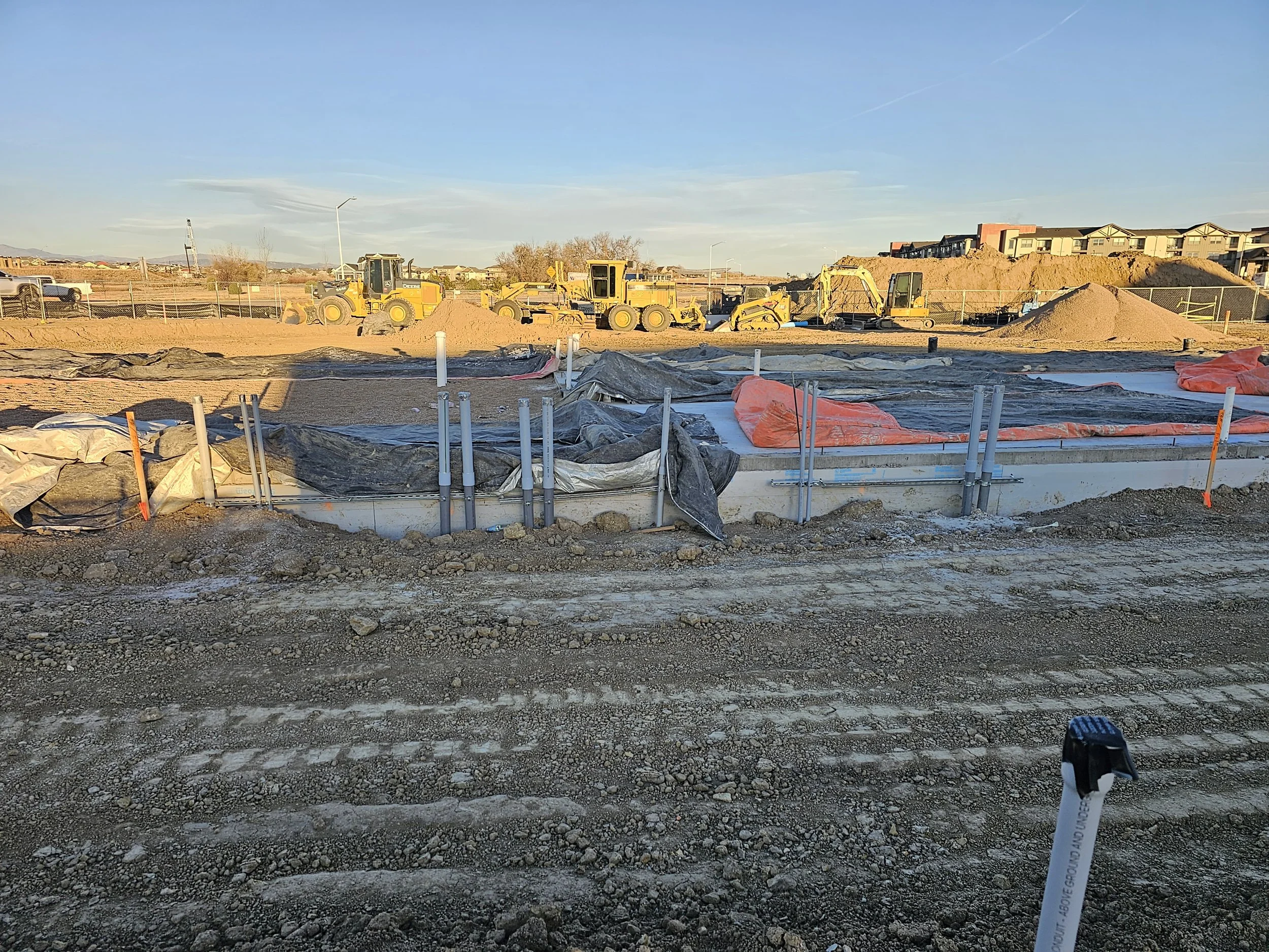 Construction site with yellow bulldozers and excavators, dirt piles, and construction materials, fencing, and residential buildings in the background under a clear blue sky.