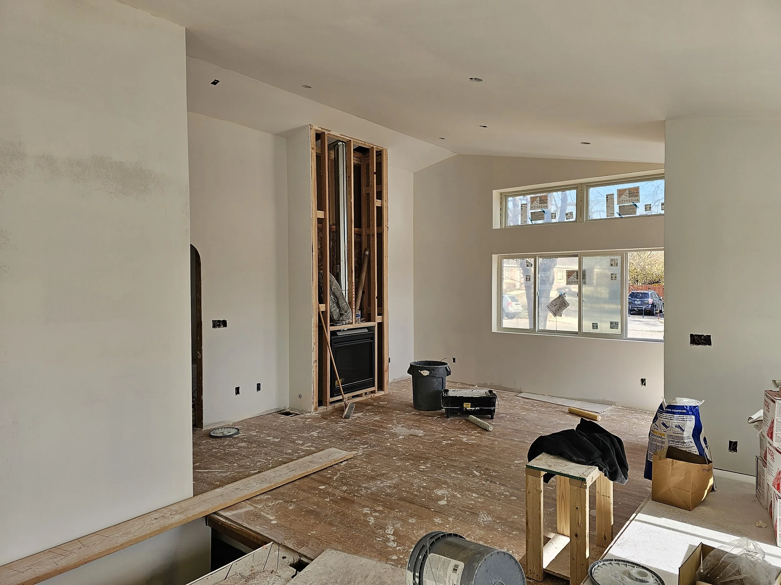 Interior of a home under renovation, showing partially finished walls, construction materials, tools, and a large window letting in natural light.