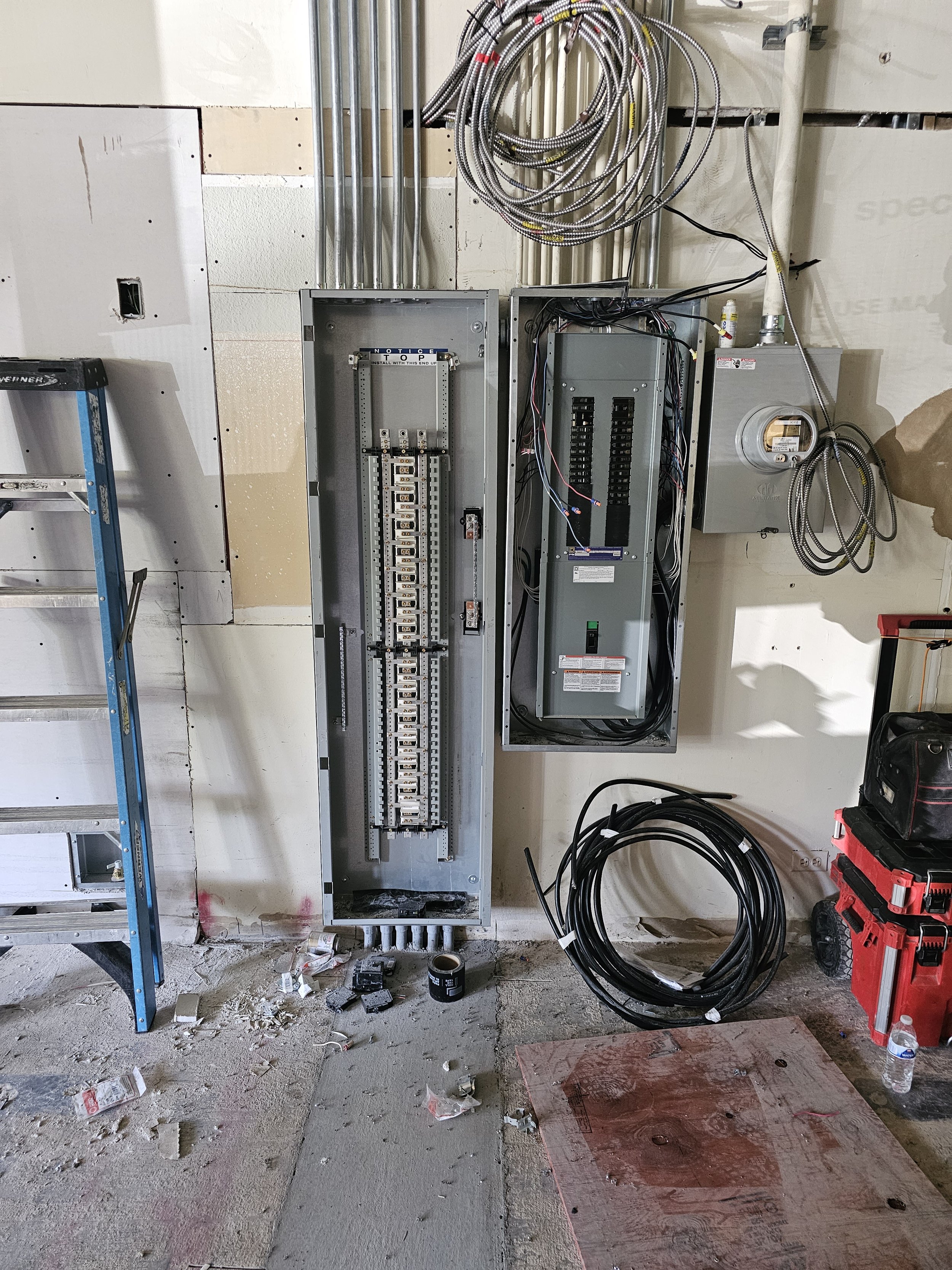 Electrical wiring and breaker panels on a construction site wall, with tools and debris on the unfinished concrete floor.