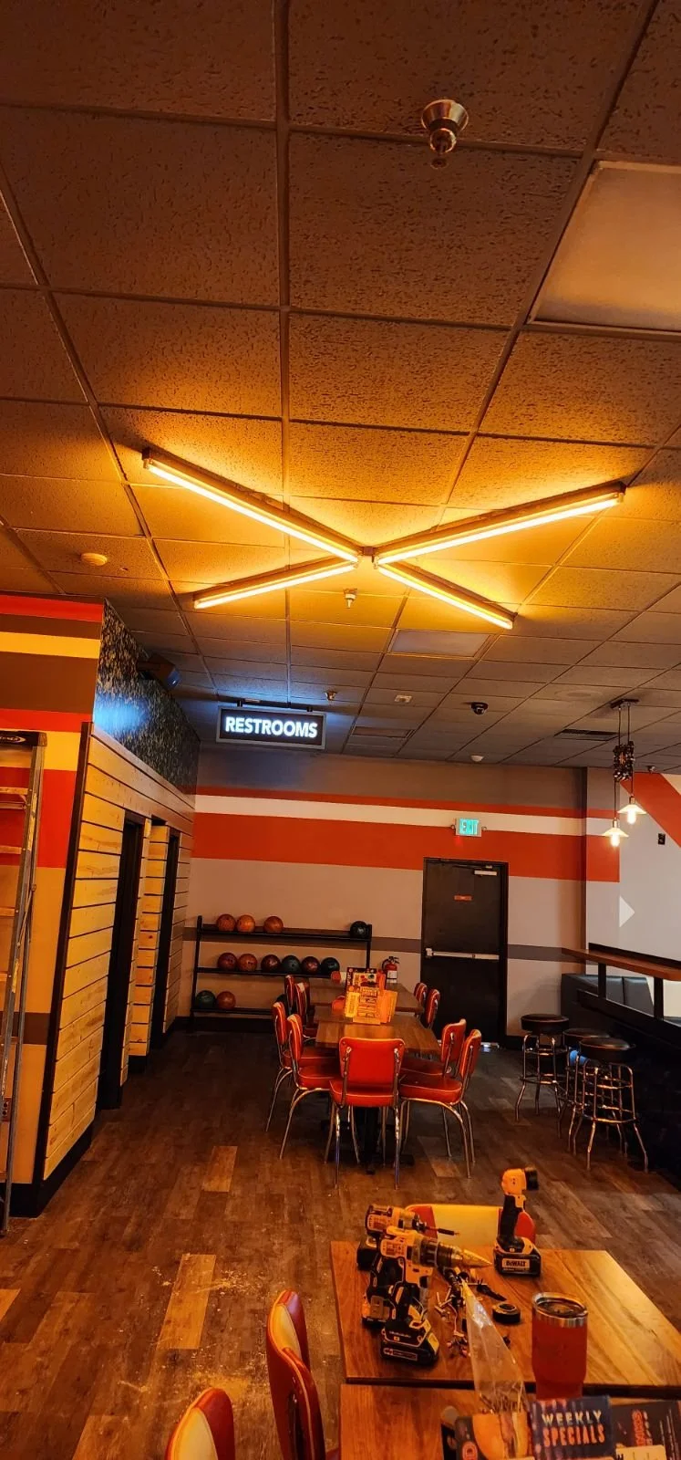 Interior of a bowling alley with a sign indicating restrooms. Bowling balls are organized on a rack, and there are tables and chairs for players.