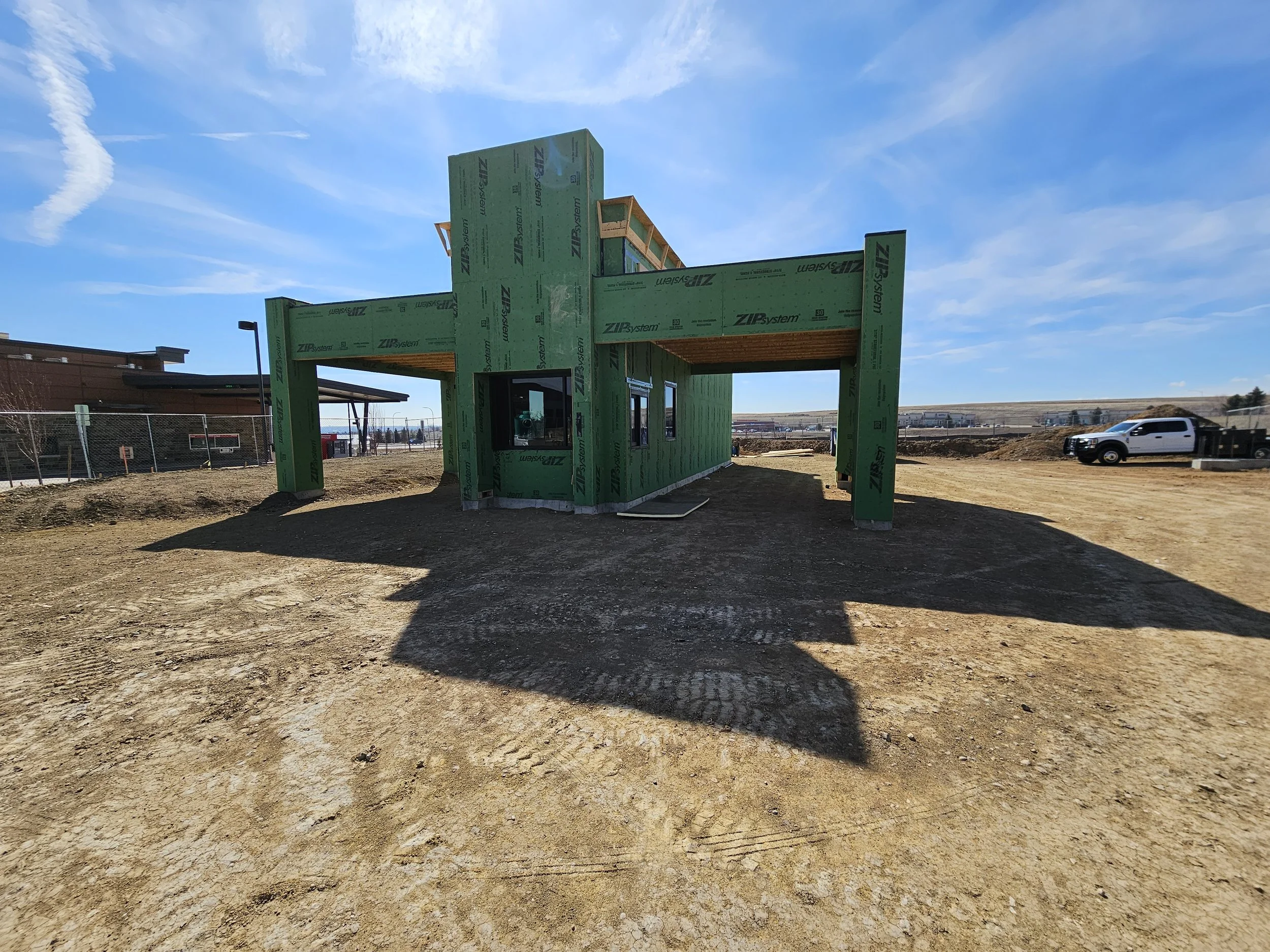 A building under construction with green insulation panels, wooden framing, and a flat roof, situated on a dirt lot with a car and distant buildings and hills in the background.