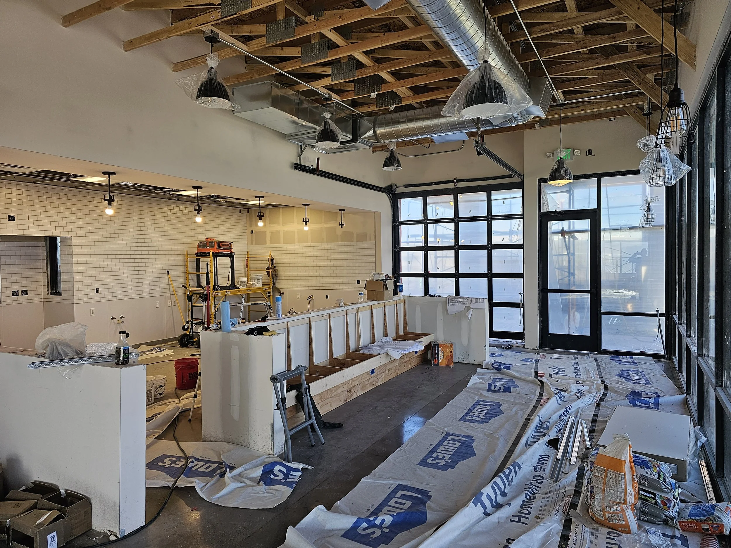 Interior of a building under construction with exposed wooden ceiling beams, ventilation ducts, light fixtures, and construction materials scattered on the floor.