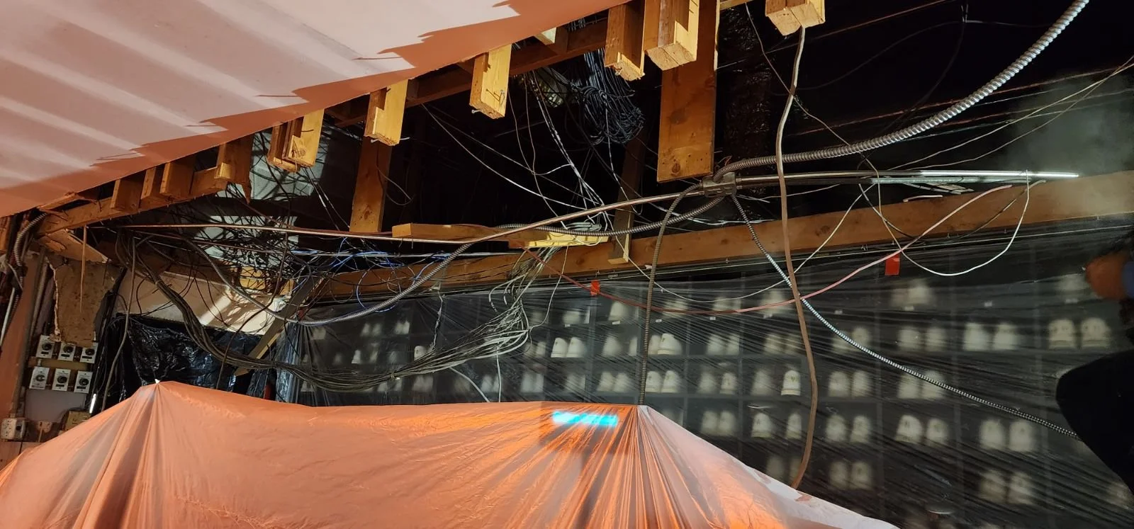 Construction site with exposed wooden beams, electrical wires, and plastic sheeting, viewed from below.