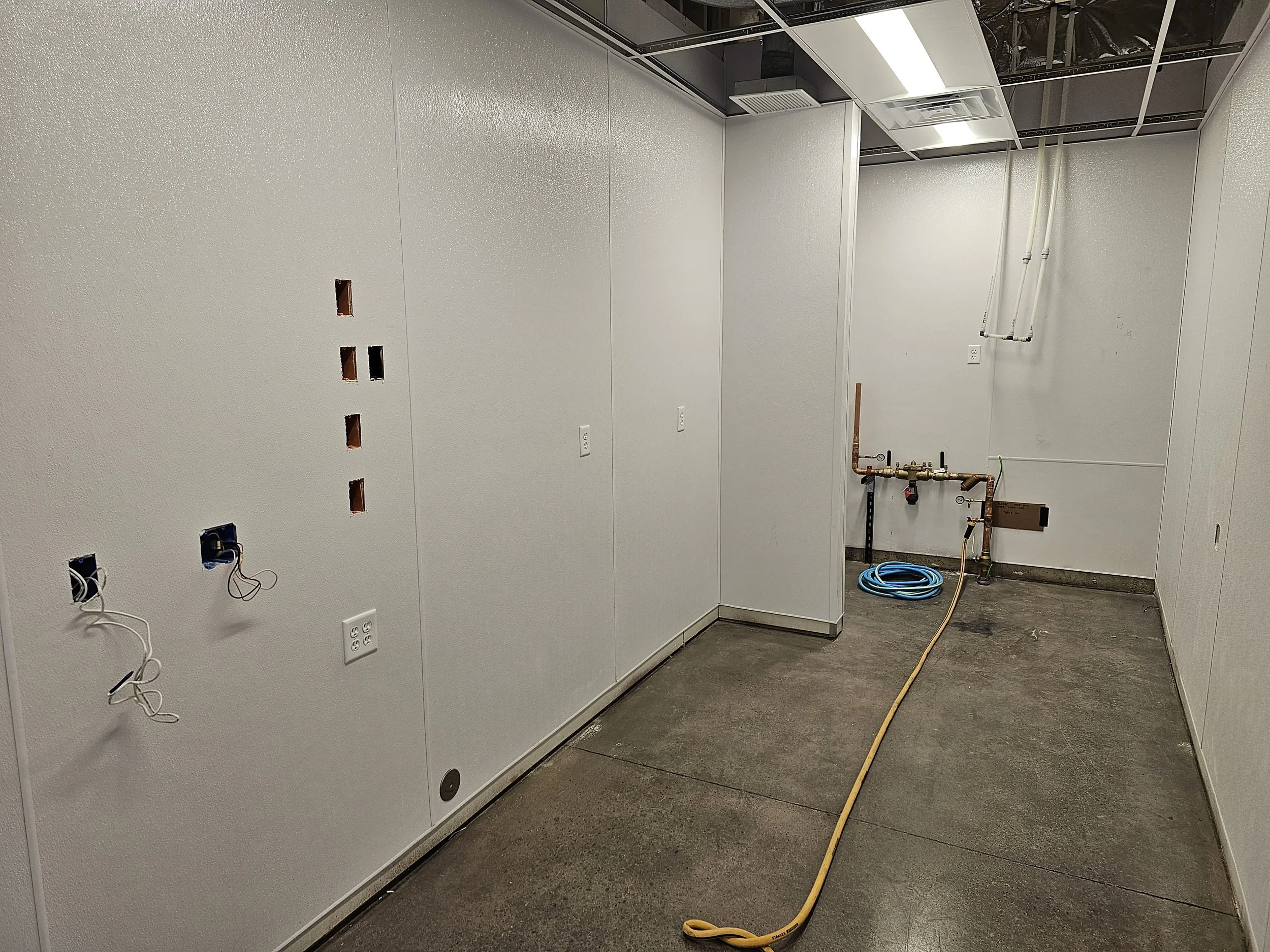 Empty room under construction with white walls, electrical outlets, and openings for electrical wiring. Exposed pipes and ventilation system on the ceiling, with a yellow extension cord on the floor.