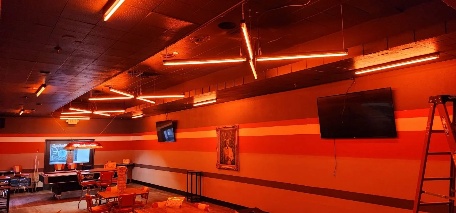 An indoor space with orange and red striped walls, a wooden ceiling, and modern orange lighting fixtures hanging from the ceiling. The room has several chairs and tables, wall-mounted TVs, a decorative picture, and a bar area with a neon sign.