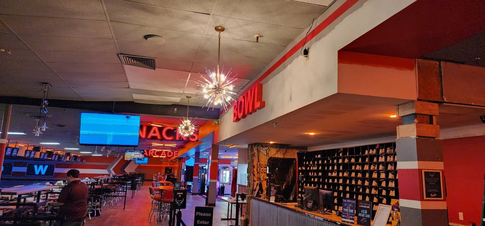 Interior of a modern bowling alley with lanes, screens, and a checkout counter, illuminated by decorative light fixtures and neon signs.