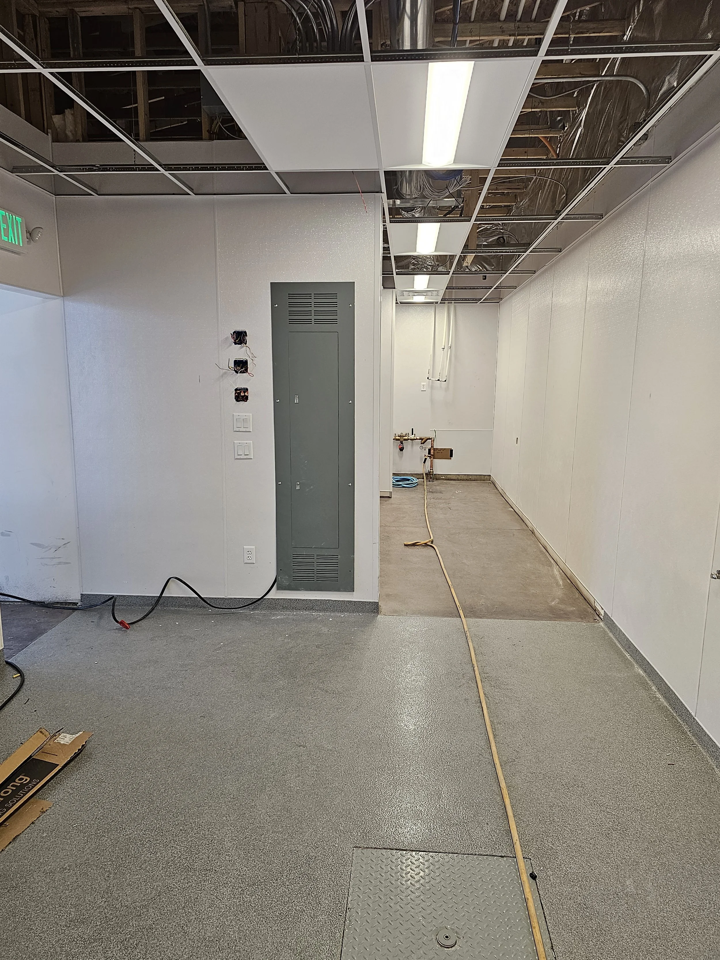 An unfinished room with a drop ceiling in progress, exposed wires, a gray electrical panel, and a yellow extension cord on the gray speckled floor.