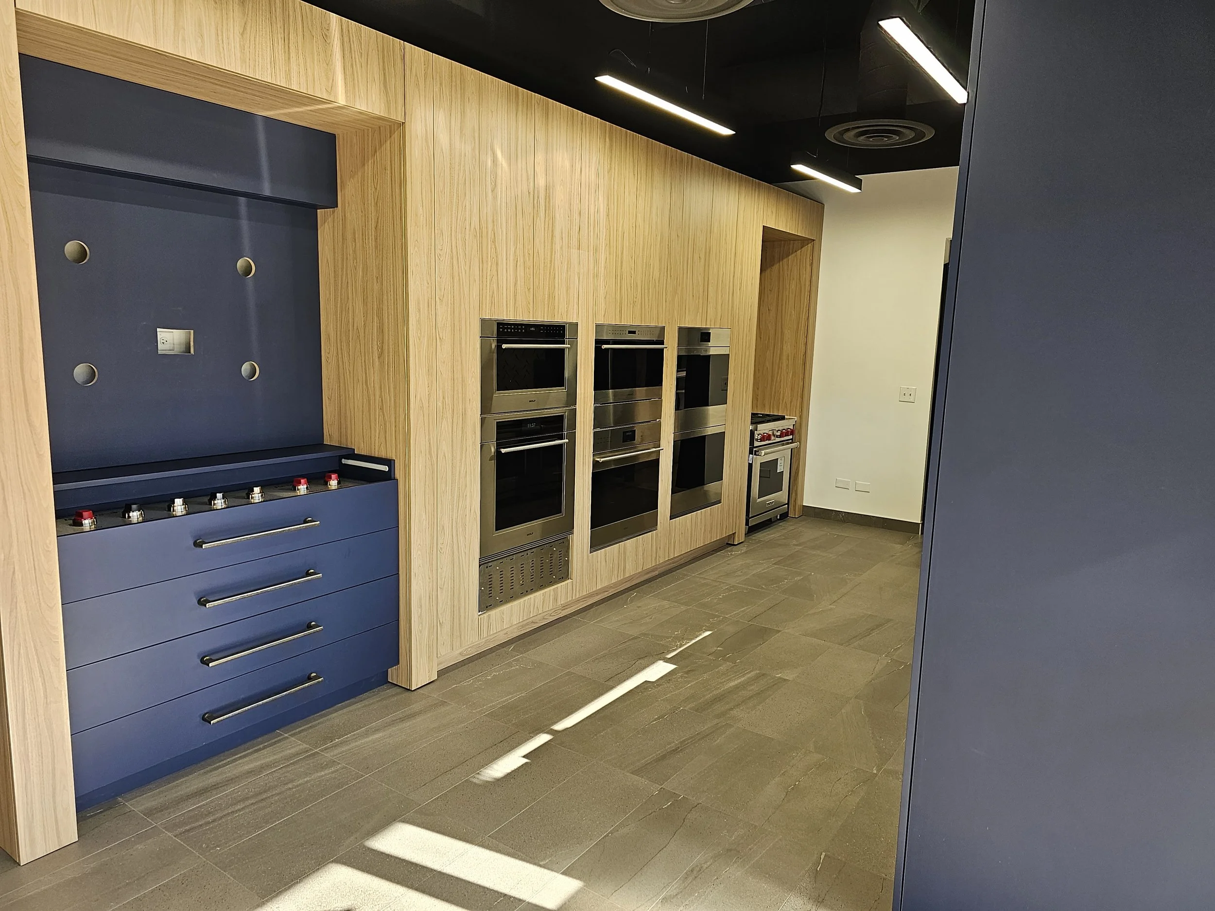 A modern kitchen wall with built-in appliances including an oven, microwave, and grill, surrounded by wooden paneling and cabinetry, with a black ceiling featuring sleek lighting fixtures.