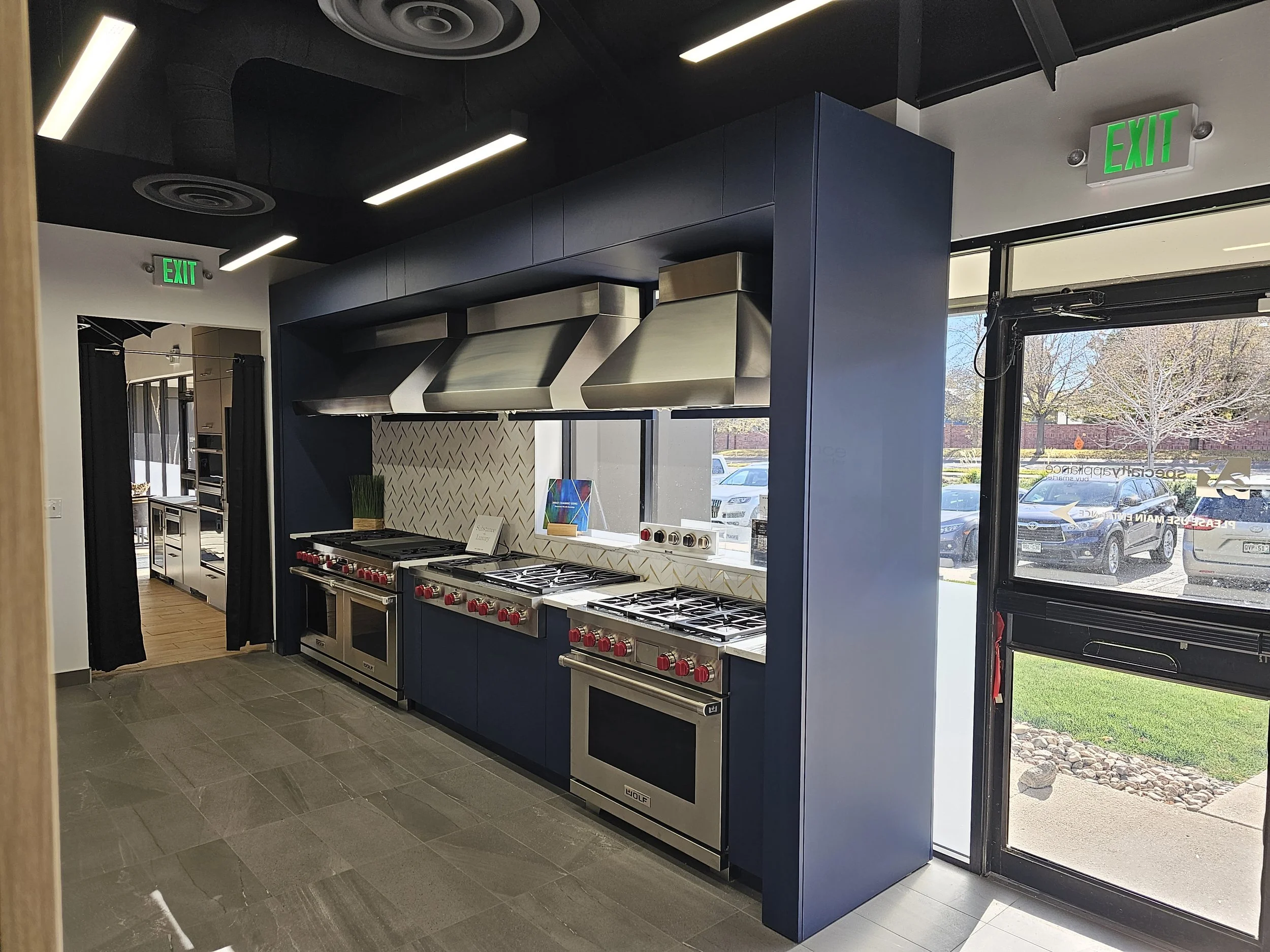 Interior of a modern showroom or store kitchen appliance display area with professional stoves and ovens, stainless steel range hoods, blue cabinetry, and large windows showing parked cars and trees outside.