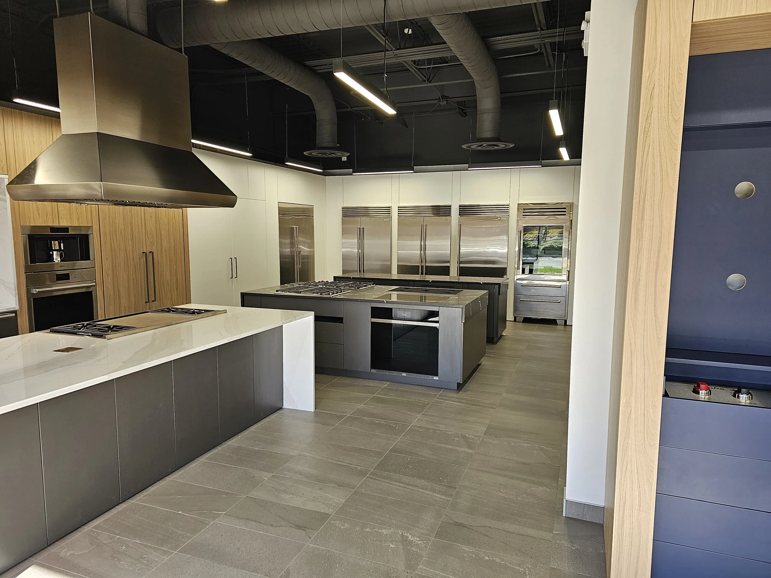 Modern kitchen with wooden cabinets, stainless steel appliances, black and gray countertops, and hanging linear lighting fixtures.
