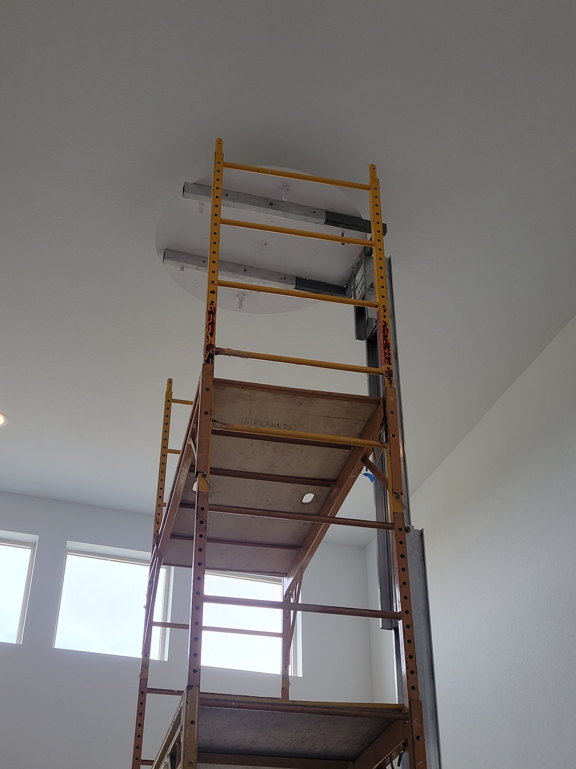 Ladder placed under ceiling with a light fixture and a drywall hole, indicating ongoing ceiling installation or repair in a room with large windows.