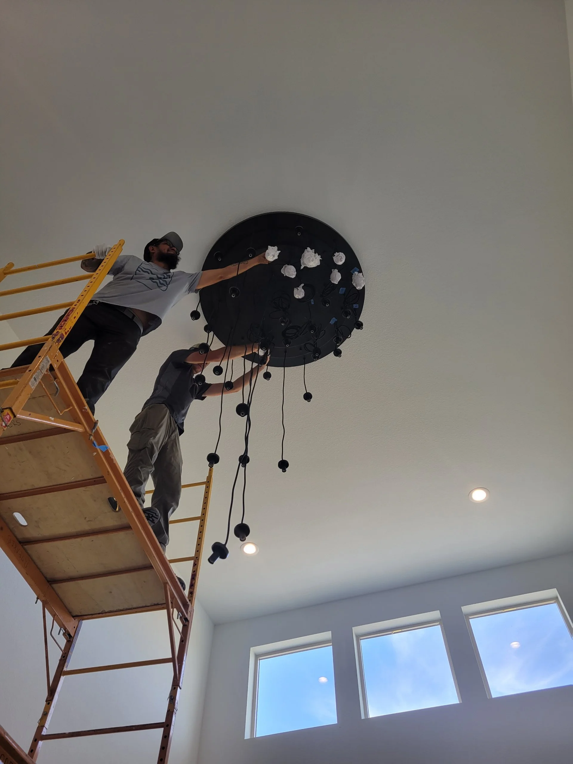 Two workers stand on a scaffold installing a black circular chandelier on a white ceiling in a room with large windows and a blue sky outside.