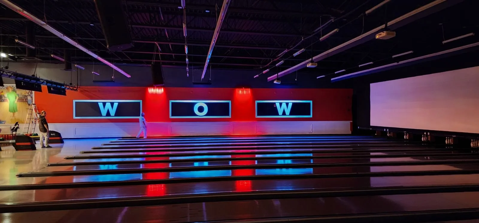 An empty bowling alley with lanes, pins, and a large screen on the right. The back wall has a red background with three large electronic displays spelling out 'W' 'O' 'W'. There are a few people walking in the alley, and the ceiling has various light