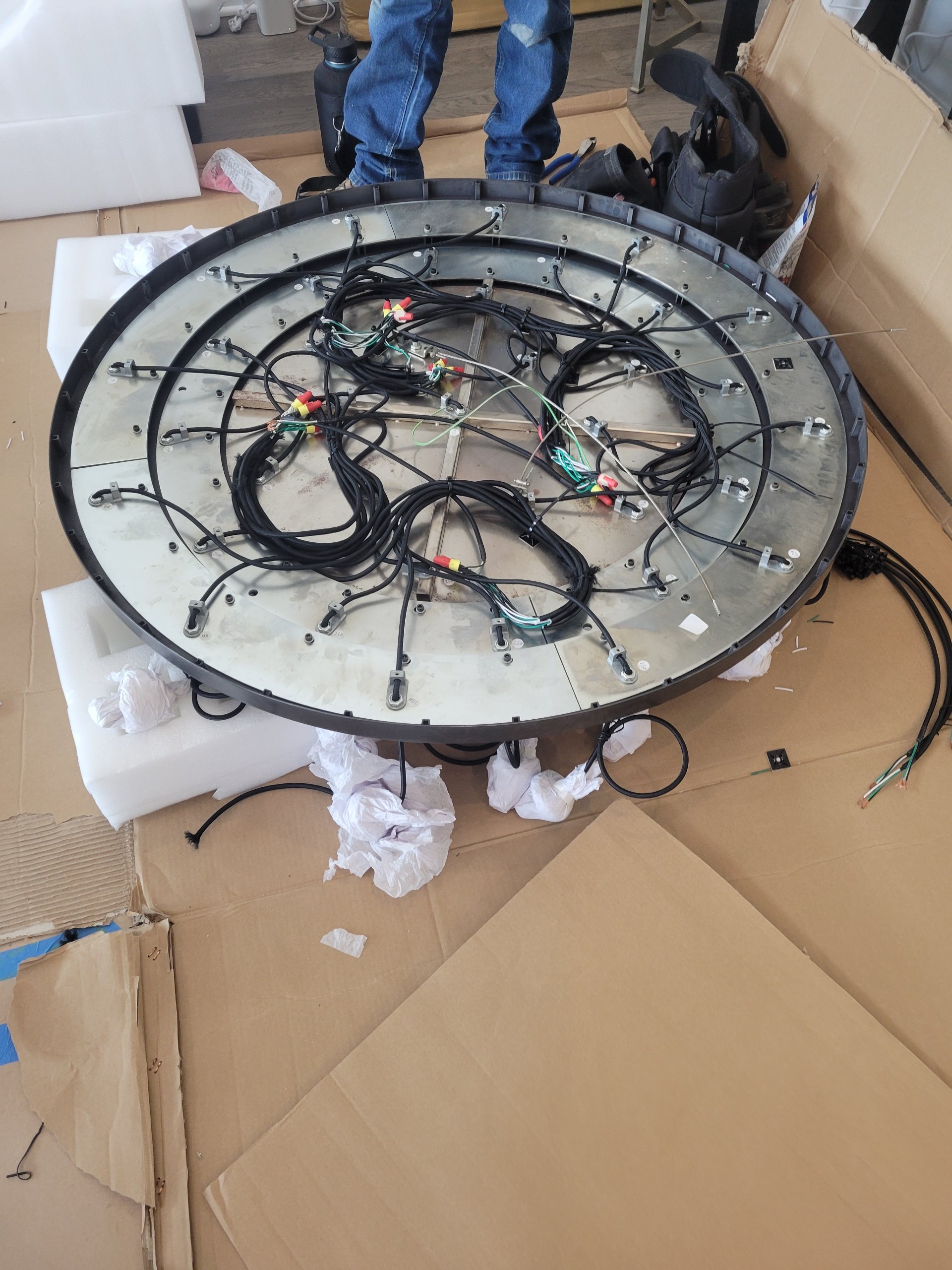 A large circular electronic device with many wires and connectors on a work surface, surrounded by cardboard boxes and tools, with a person standing behind it.