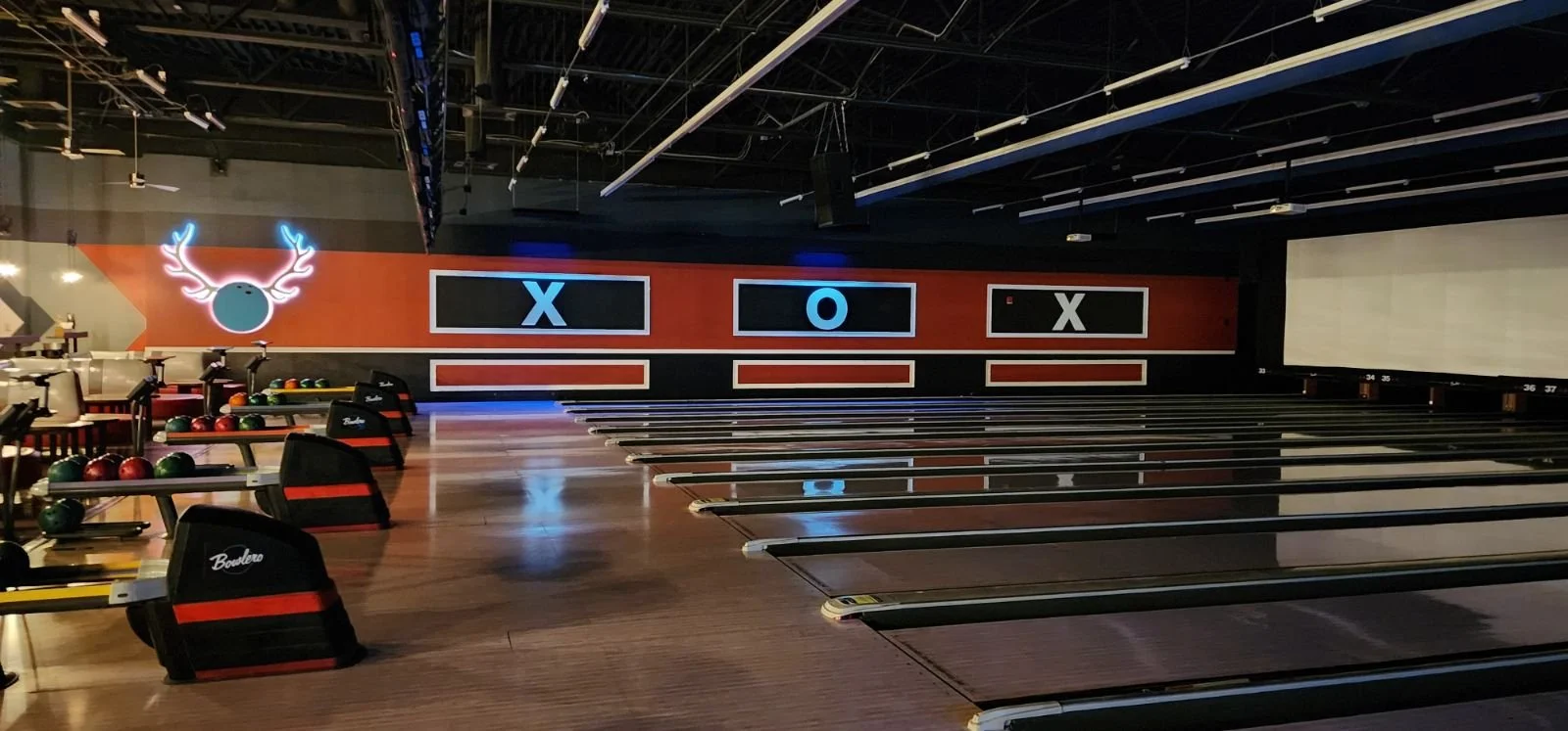Empty bowling alley with several lanes, bowling balls on racks, a neon reindeer sign, and a wall with 'X' and 'O' game screens.