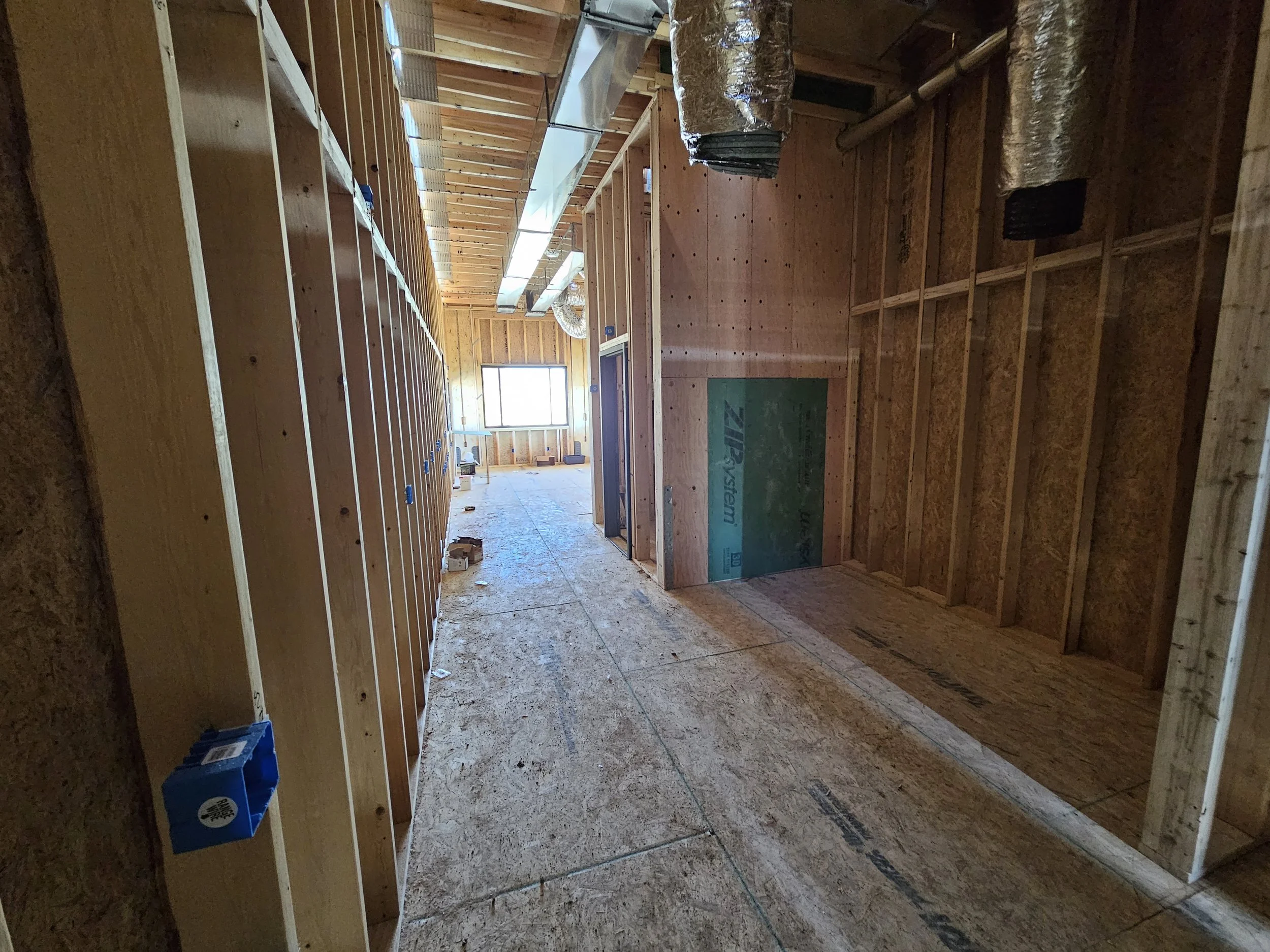 Interior view of a house under construction with wooden framing on walls and ceiling, plywood floor, ductwork on ceiling, electrical outlets on walls, and a window at the far end letting in natural light.