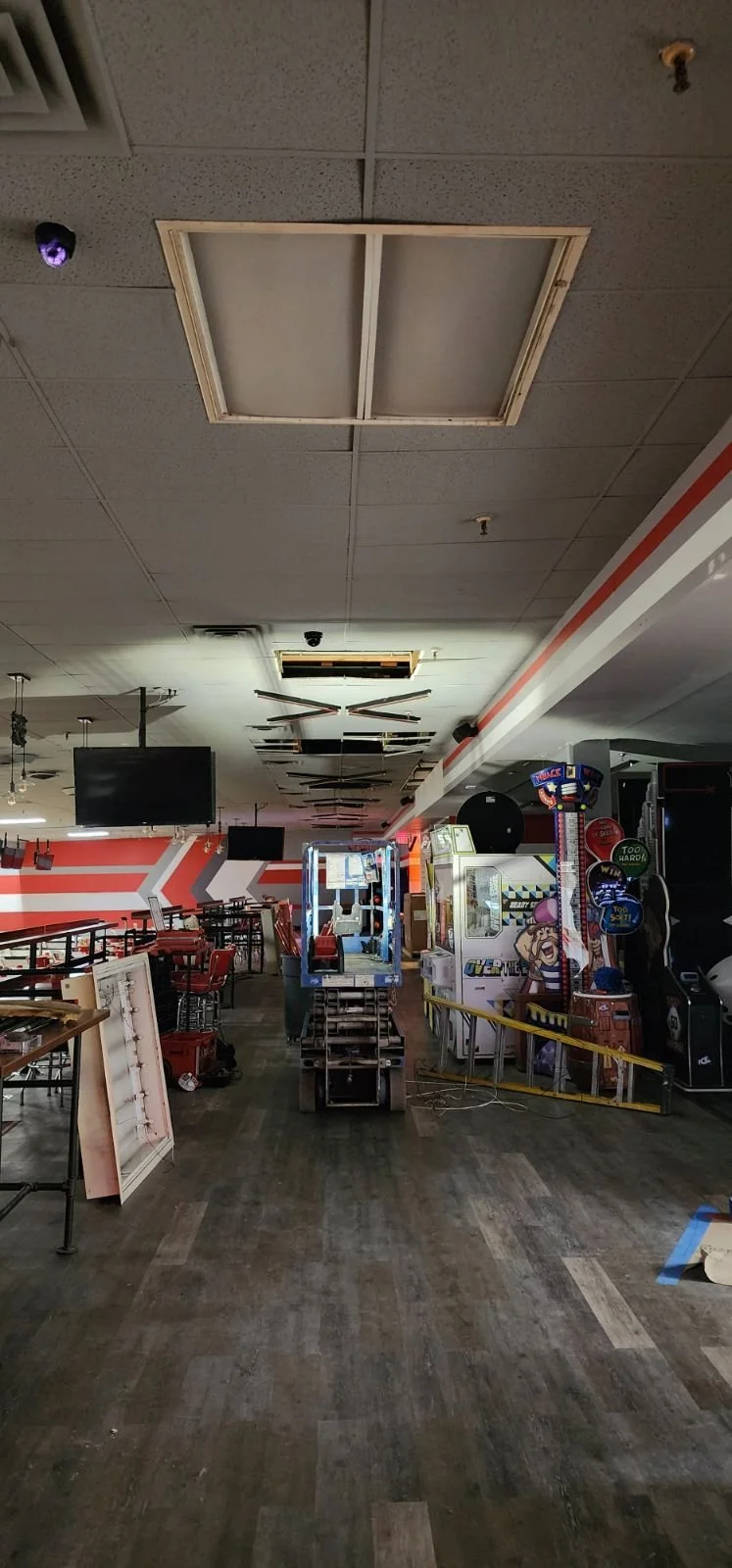 An arcade with missing ceiling tiles, some damaged or removed, and a partially disassembled climbing game machine. The floor is wooden, and there are red chairs and tables. The backdrop has a red and white striped wall with some decorative arrow patt