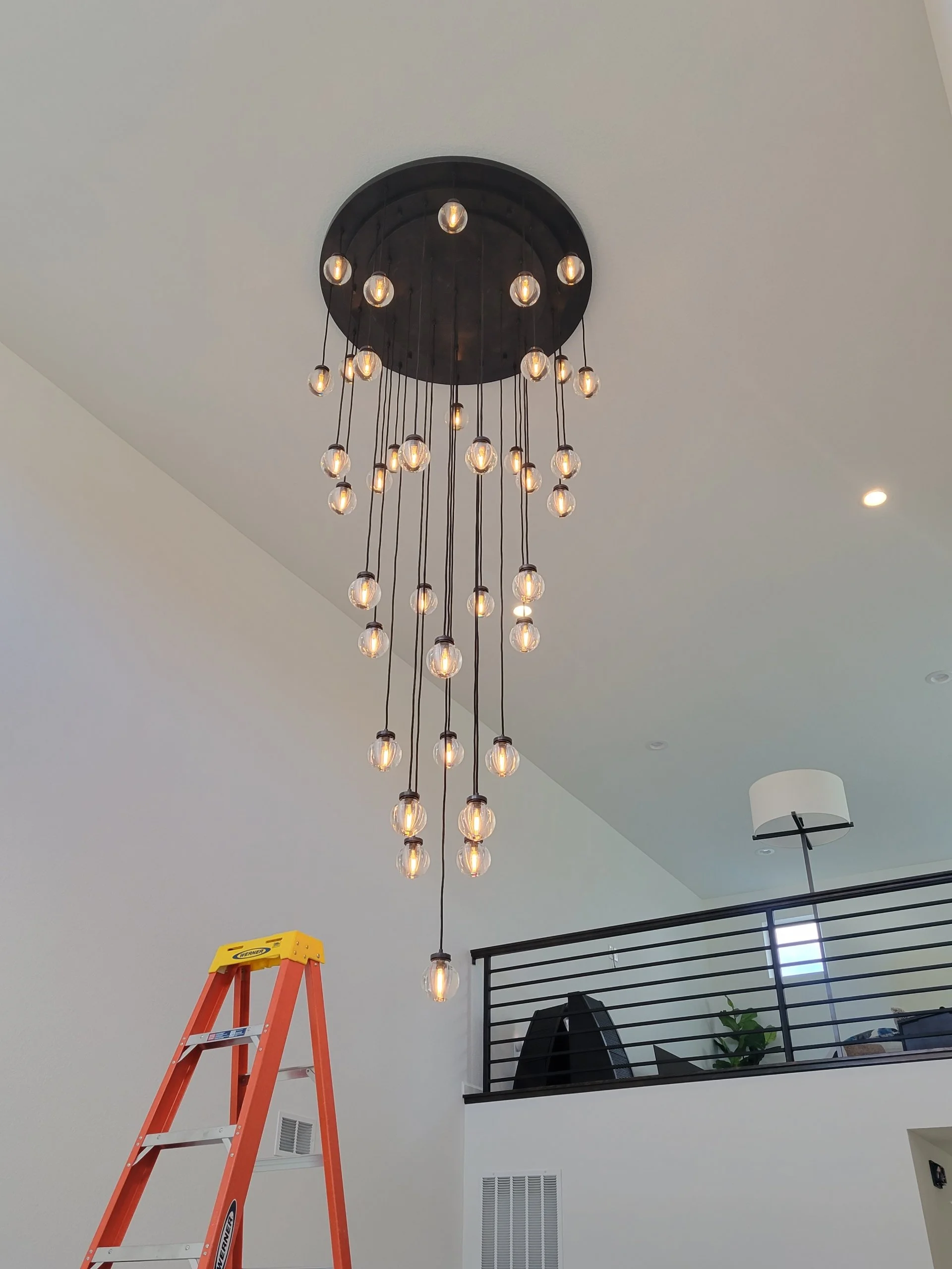 Interior of a modern home with a stylish chandelier hanging from the ceiling, an orange ladder in the foreground, and a black rail balcony with a potted plant in the background.