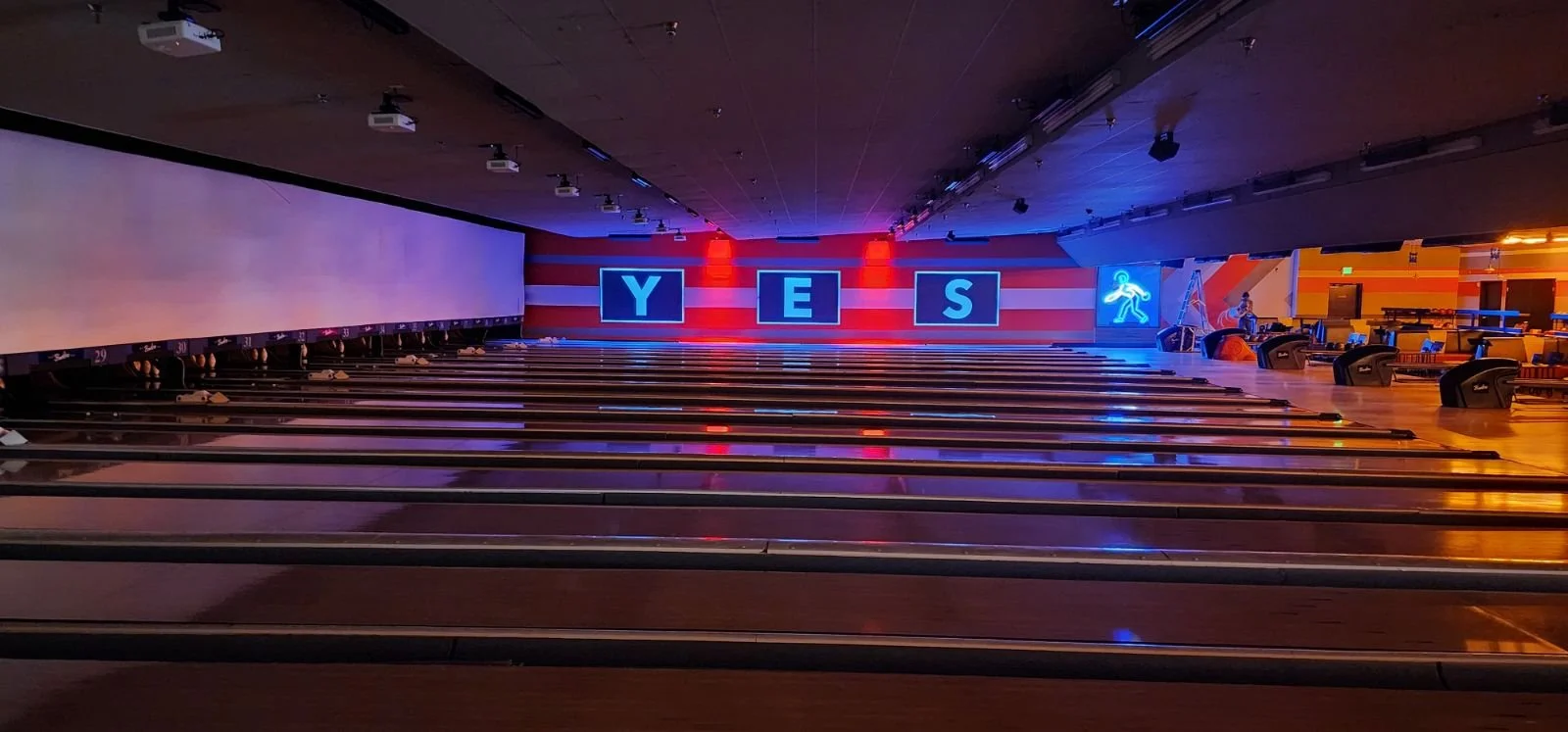 An empty bowling alley with lanes and pins visible, illuminated by colorful neon lights, including a large neon 'YES' sign on the back wall.
