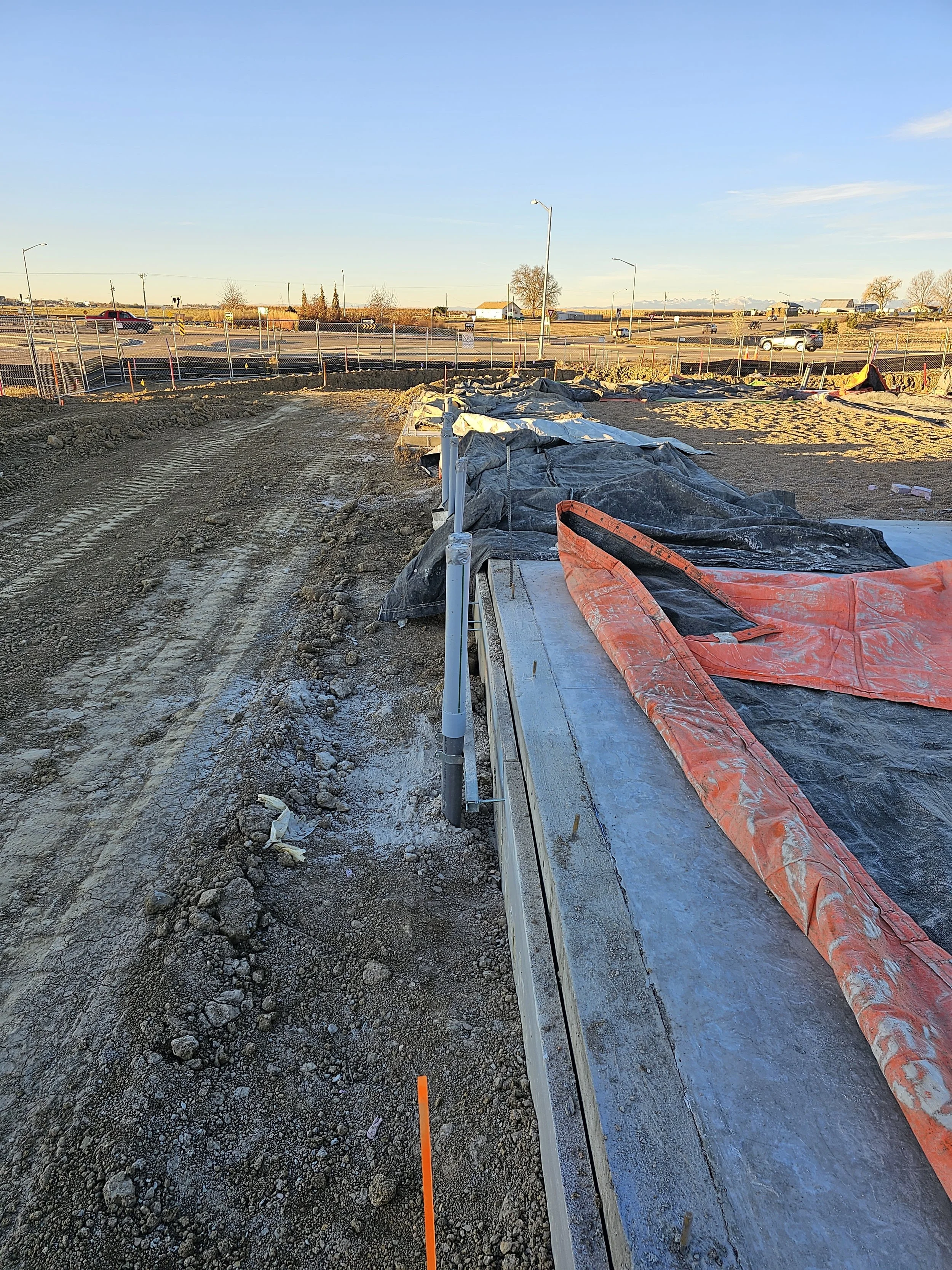 Construction site with dirt and gravel ground, covered with black and orange tarps, in a developing area with fencing, street lights, and cars in the background, under a clear blue sky.