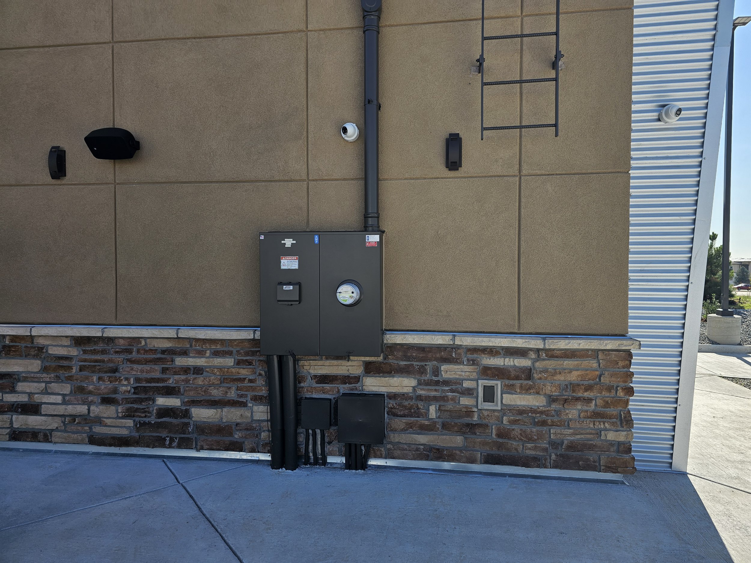 Exterior wall of a building with electrical and security equipment including a black speaker, two camera lenses, a black box, and a ladder, against beige wall with stone at bottom and a parking lot in background.