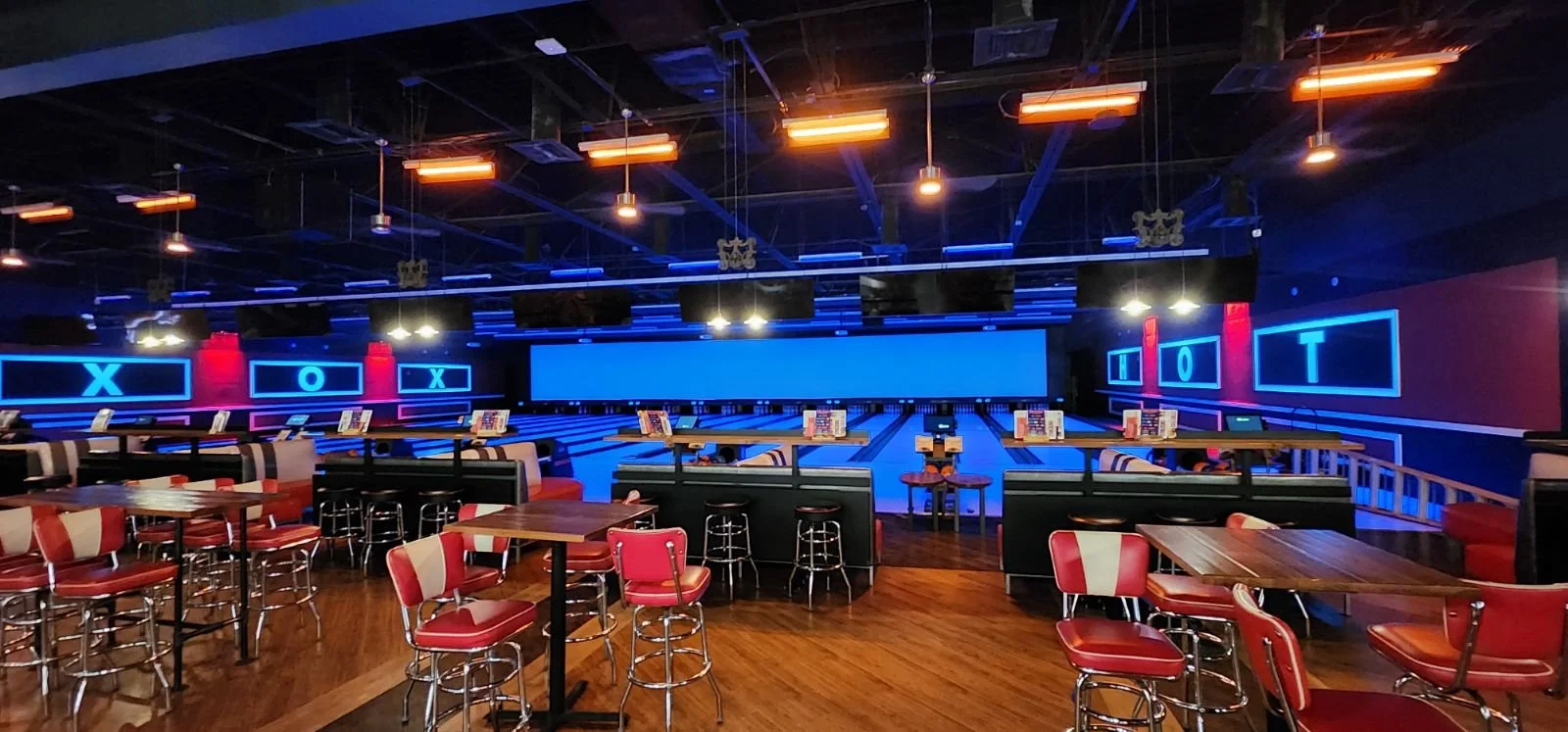 Indoor bowling alley with multiple lanes, glowing neon blue and red lights, and seating areas with red and beige chairs.